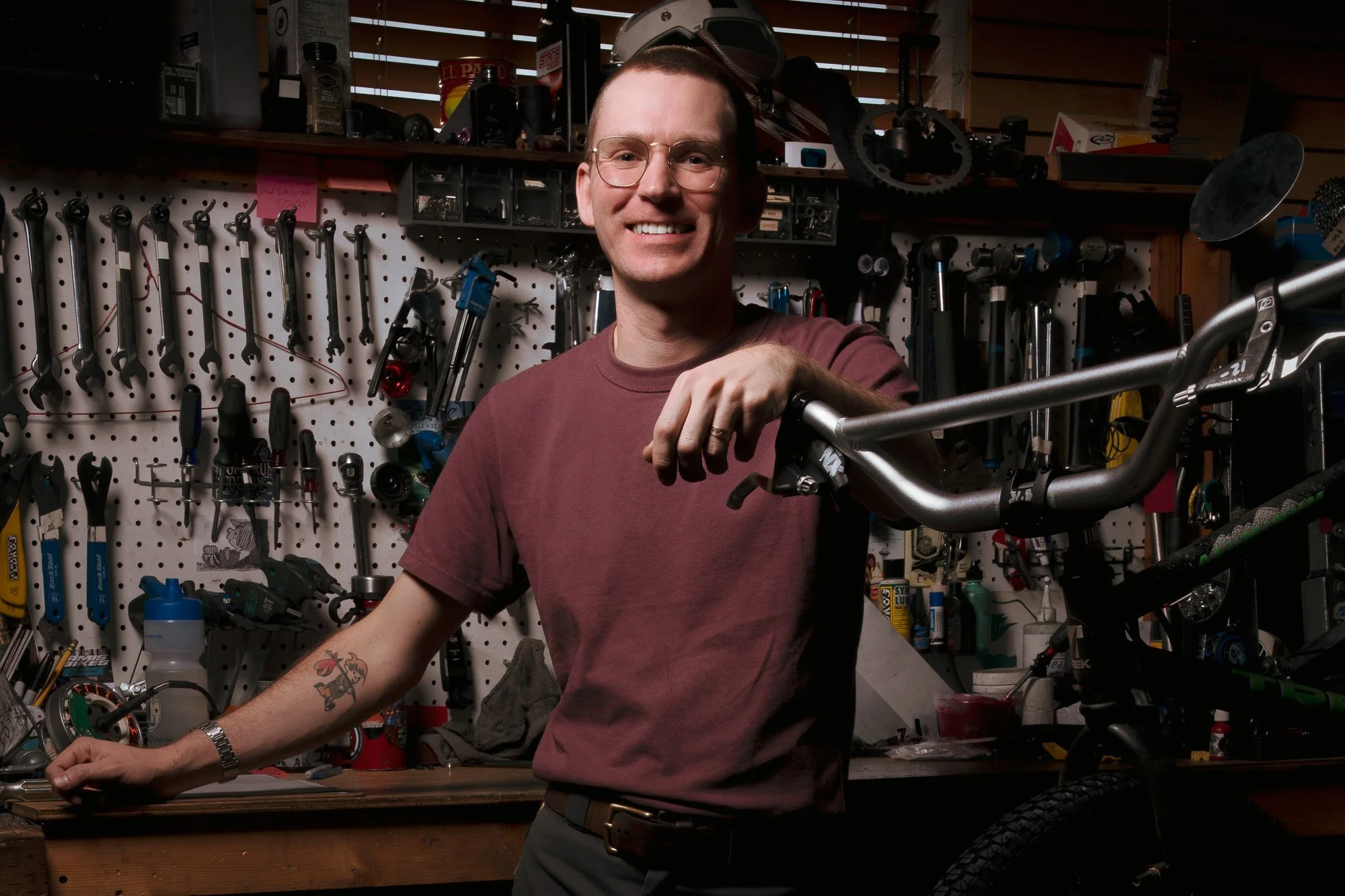 A smiling man with glasses and a tattoo on his arm in a workshop, surrounded by various tools and bicycle parts, holding a bicycle handlebar.