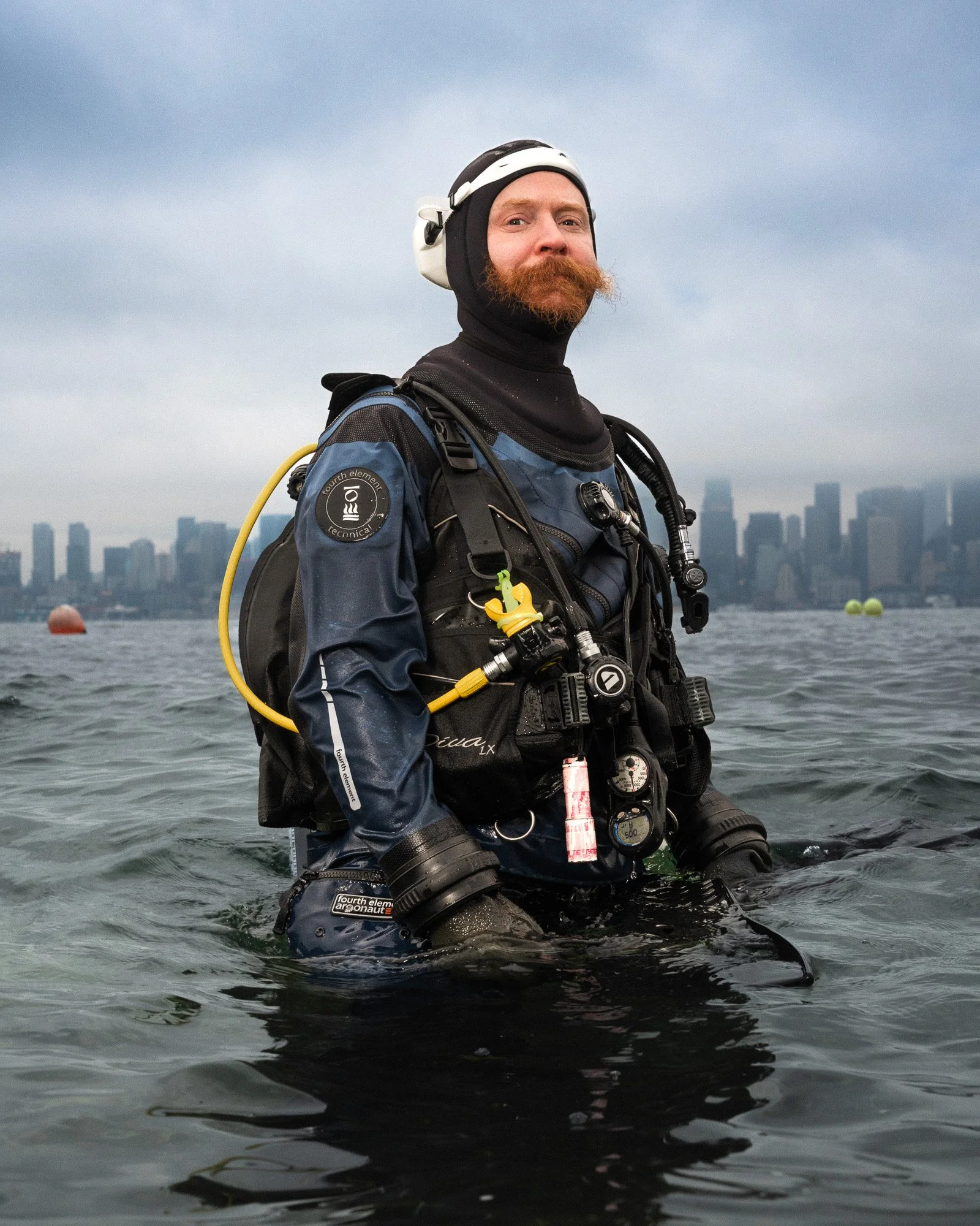 A man with a red beard and mustache wearing a black and white helmet and a wetsuit, standing in water with a cityscape in the background, possibly ready for diving or underwater activities.