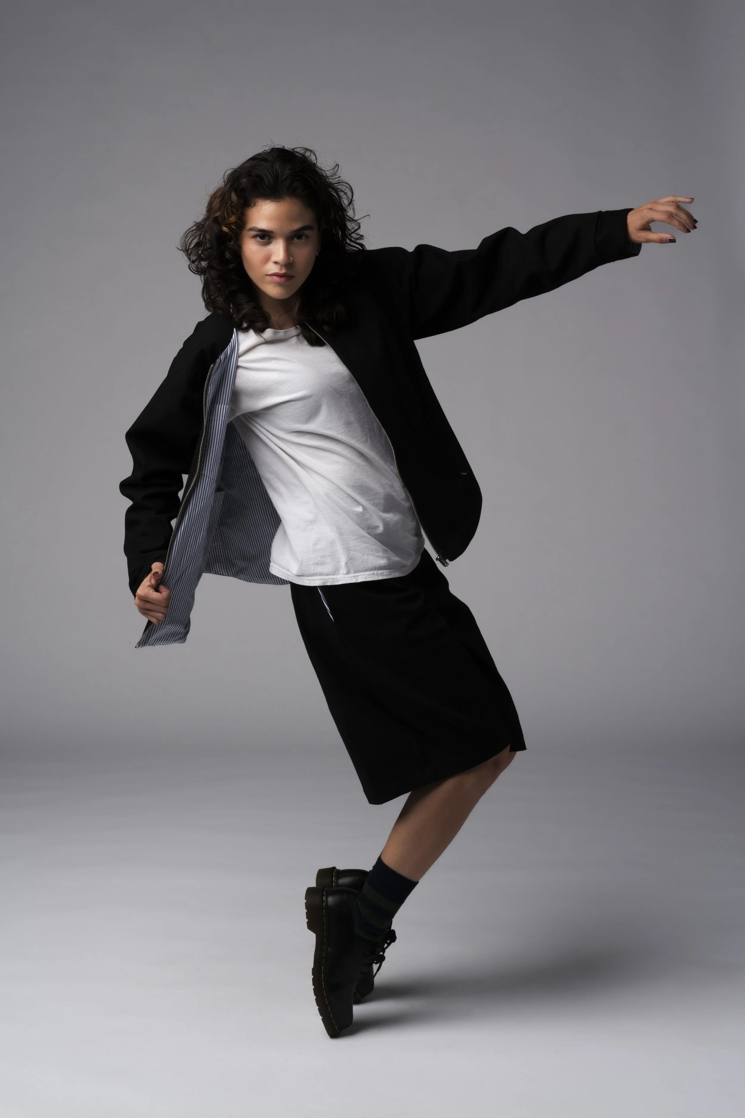 Young woman with curly hair posing in a studio against a gray background, wearing a black jacket, white t-shirt, black skirt, and black boots, balancing on one foot with one arm extended.