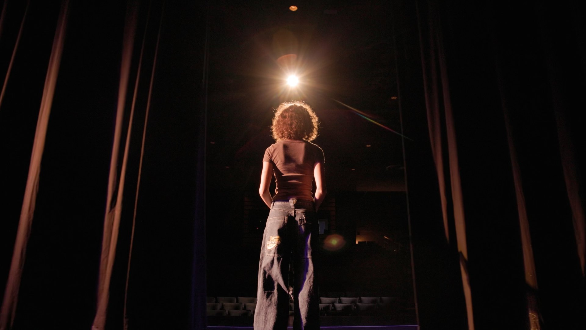 A person with curly hair standing on stage under bright lighting, viewed from behind, with theater seating visible in the background.