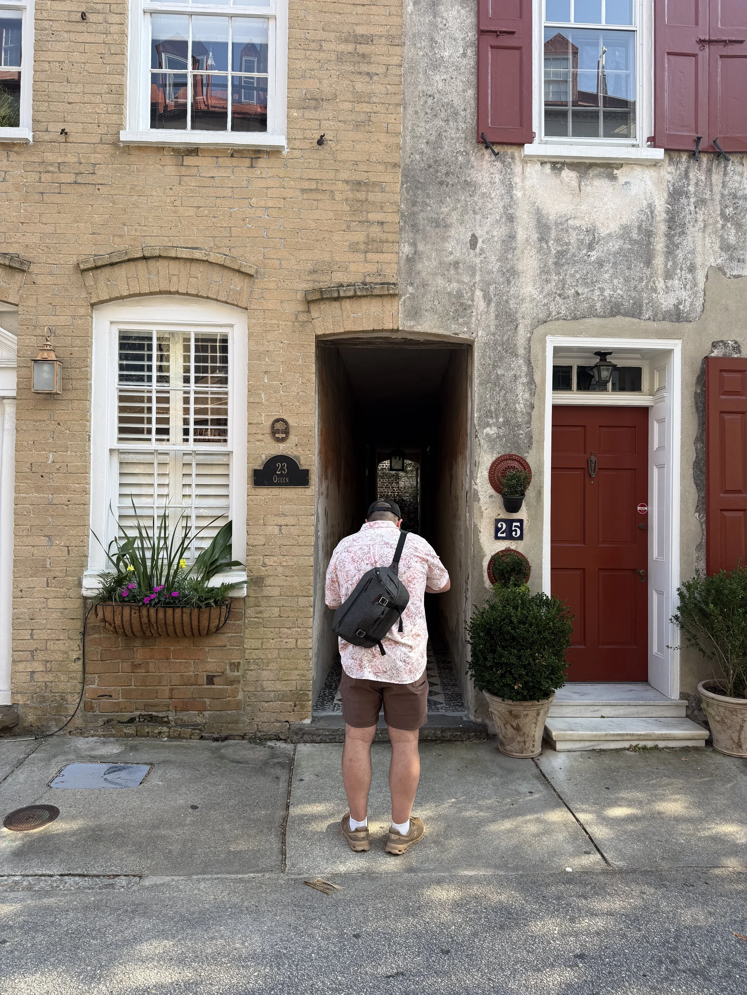 A man with a backpack and shorts is walking on the sidewalk in front of a row of connected houses. The houses have brick and concrete exteriors, with windows featuring white trim and red shutters. There are plants and decorative items near the entrance of each house. The man is facing away from the camera, and a partial view of the street is visible.