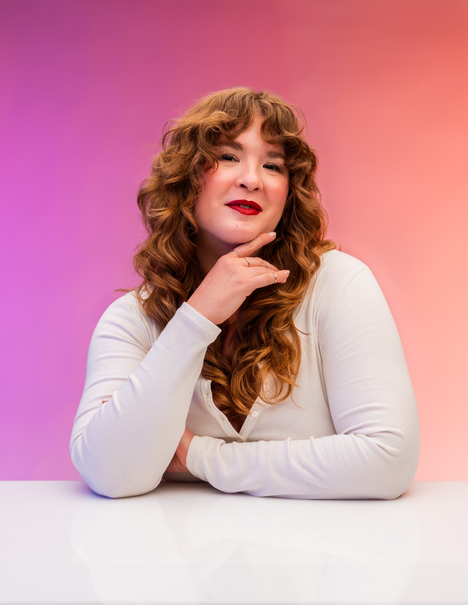 A woman with curly red hair and red lipstick sitting at a white table with a pink and orange gradient background.