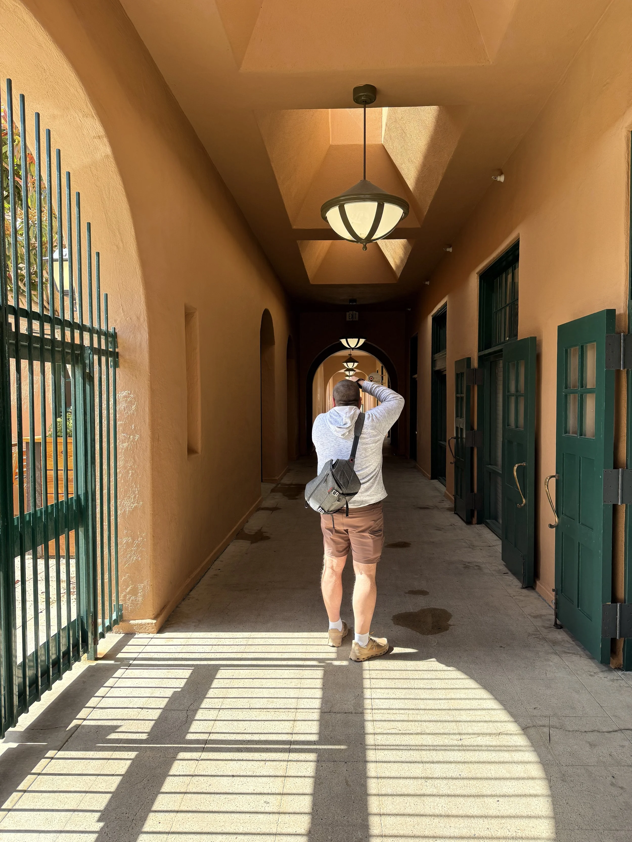 Man with short hair, wearing a white hoodie, brown shorts, and tan shoes, taking a photo with his right hand in a corridor with yellow-orange walls, green doors, and ceiling-mounted lights creating shadow patterns on the floor.