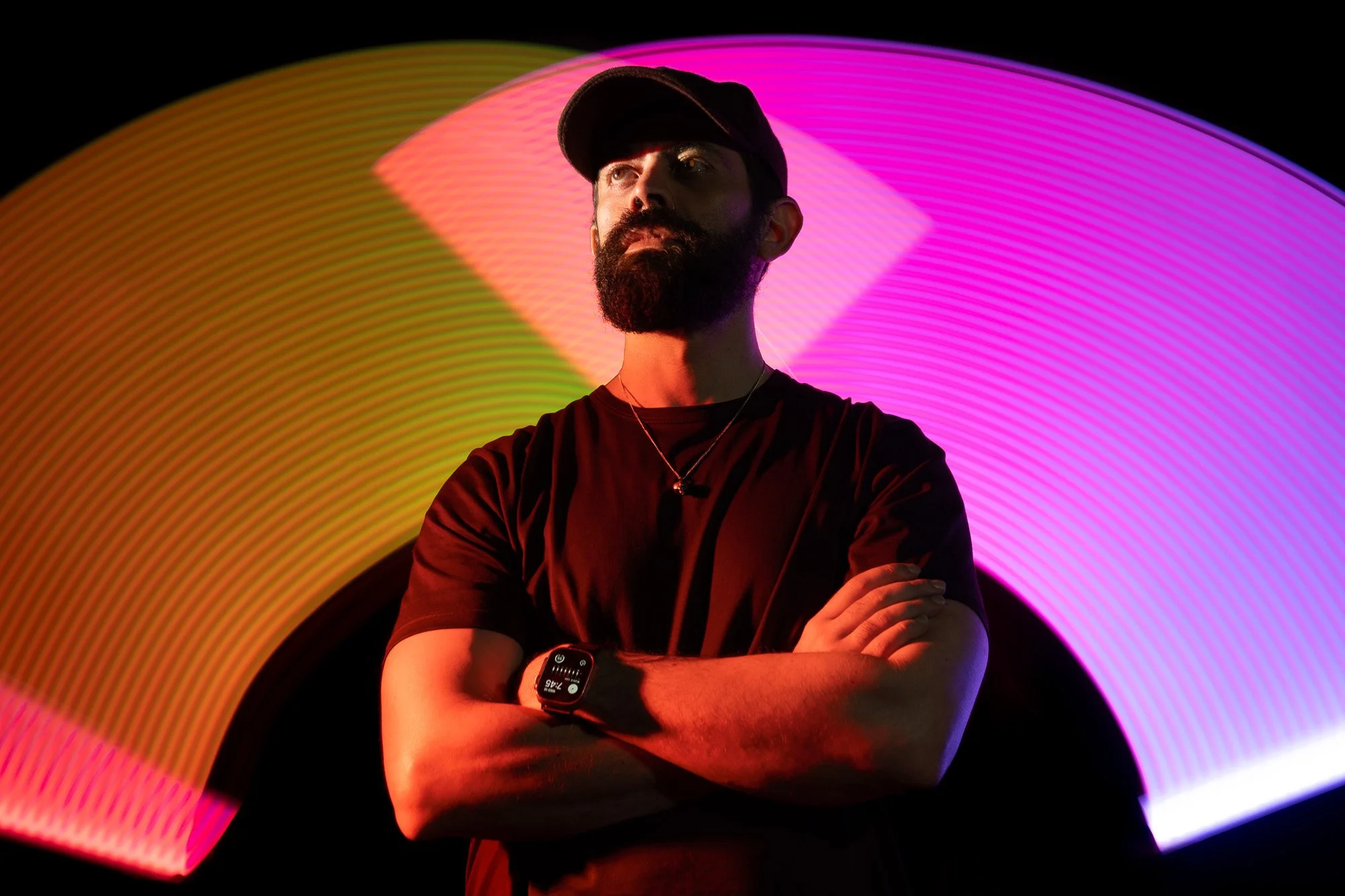 A man with a beard wearing a black cap and t-shirt, crossing his arms, standing in front of colorful light trails against a black background.