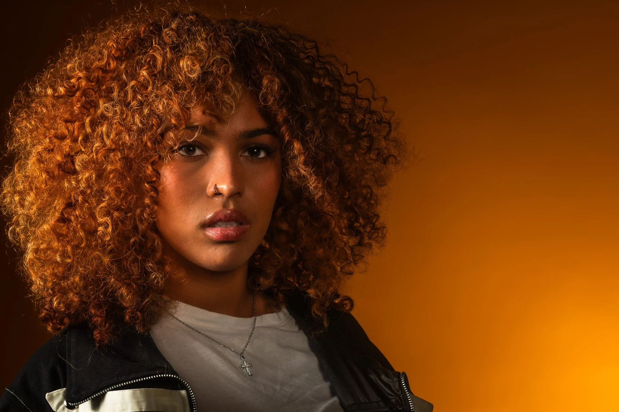 A woman with curly reddish hair, light brown skin, and a silver nose ring, wearing a white shirt, black jacket, and a cross necklace, standing against a warm brown background.