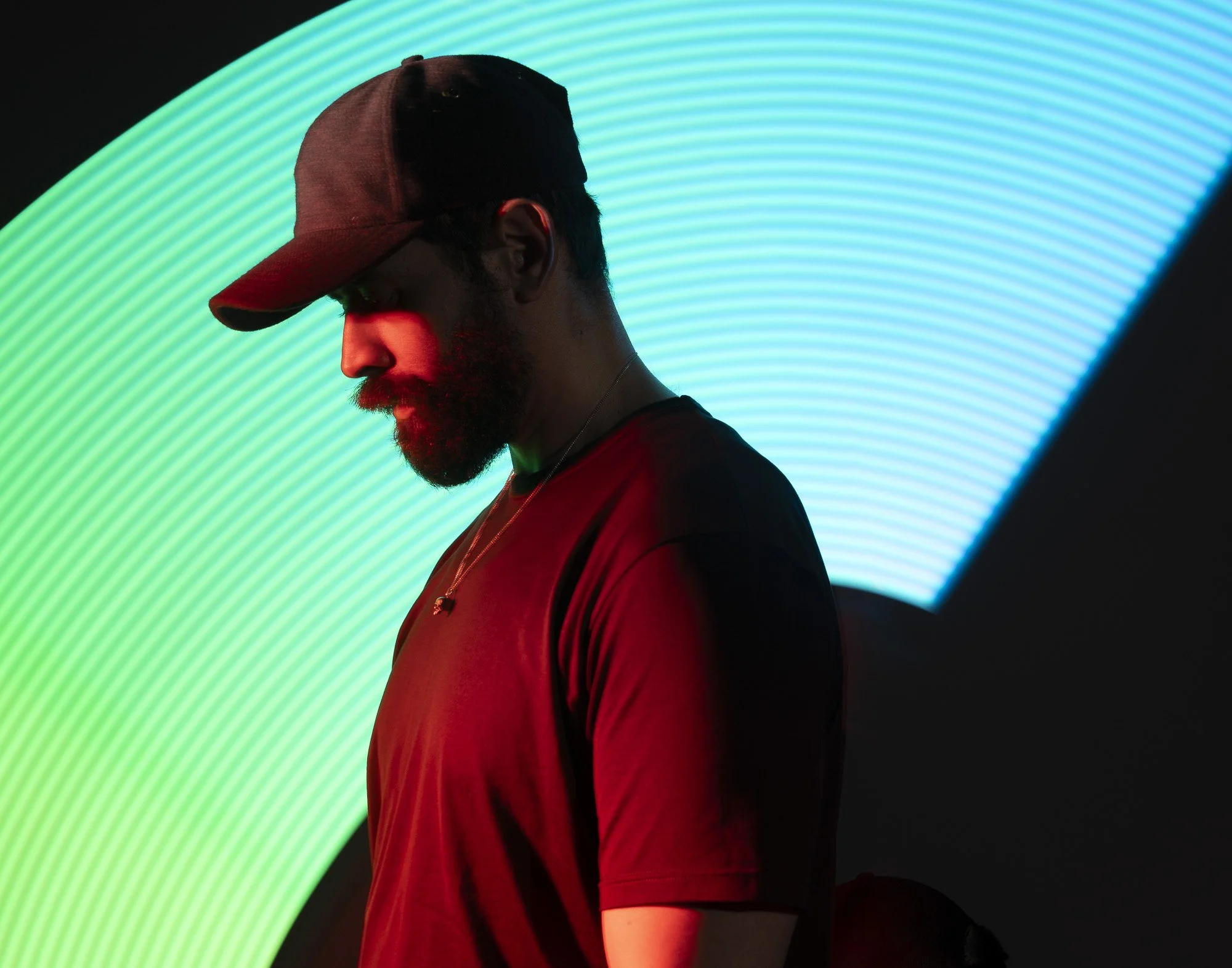 A person wearing a red cap and shirt, with a beard, is looking downward against a background of vibrant green neon light arcs.