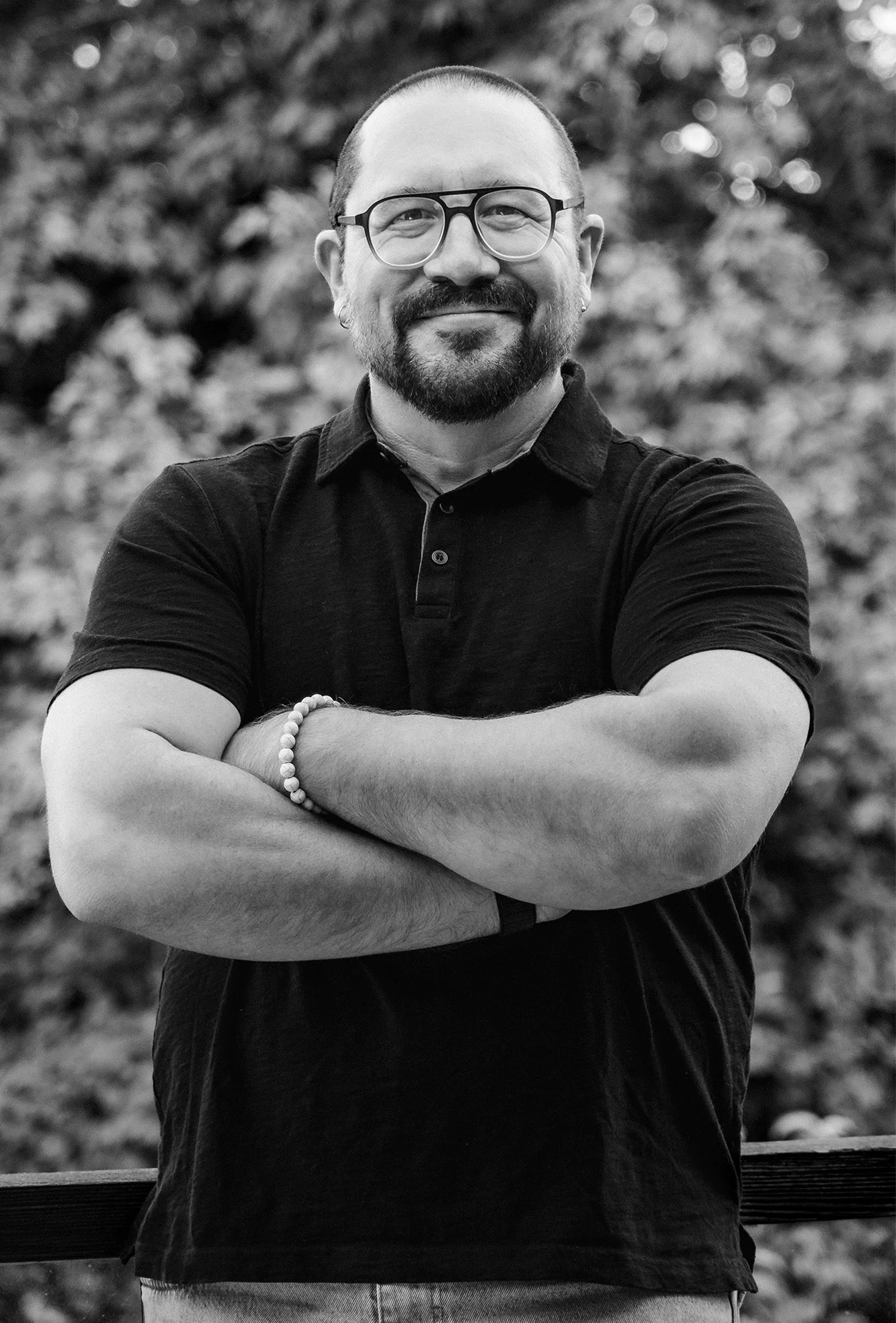 A black and white photo of a man with glasses, a goatee, and a bald head, smiling with arms crossed, standing outdoors with blurred trees in the background.