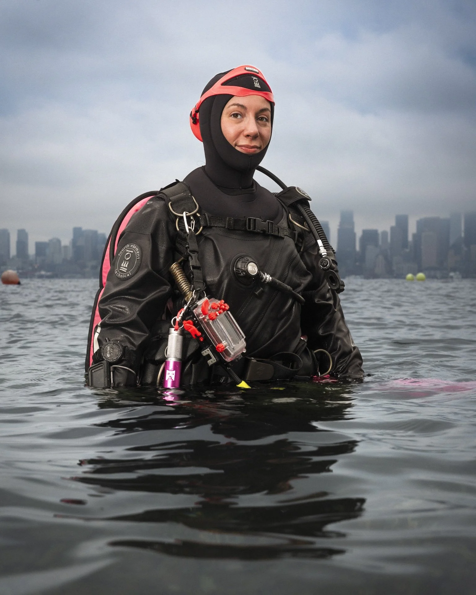 A woman in deep-sea diving gear standing in water with a city skyline in the background, overcast sky.