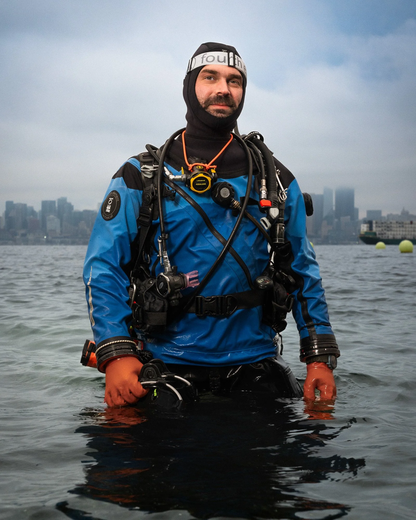 A diver standing in the water, wearing a blue wetsuit, scuba gear, and a black hood, with an overcast city skyline in the background.