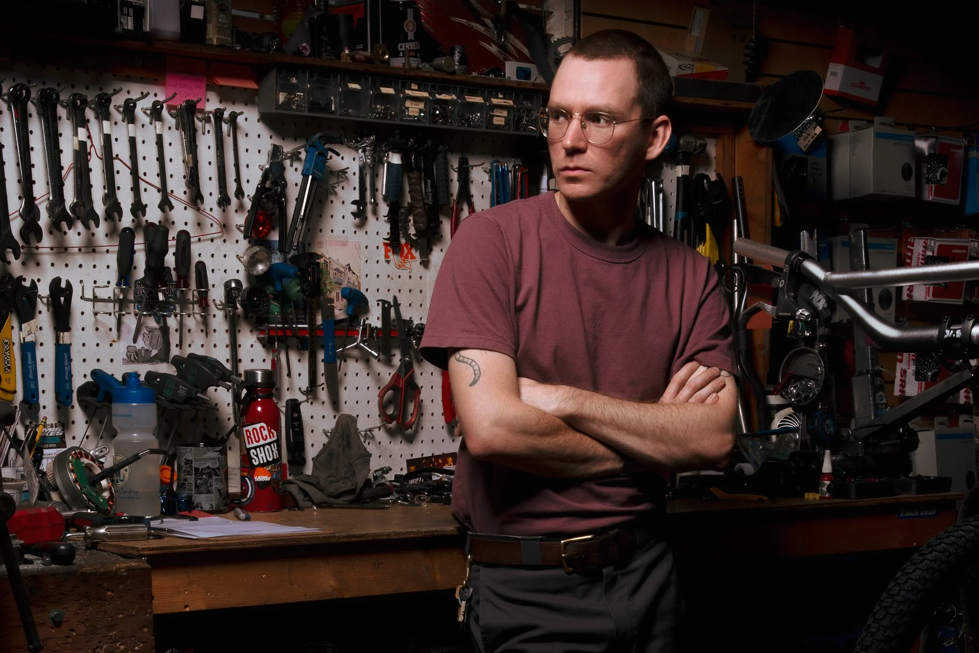 A man with short dark hair and glasses, wearing a maroon T-shirt, stands with arms crossed in a cluttered workshop or bike repair shop. The background features a white pegboard wall filled with various tools, including wrenches, screwdrivers, and oth