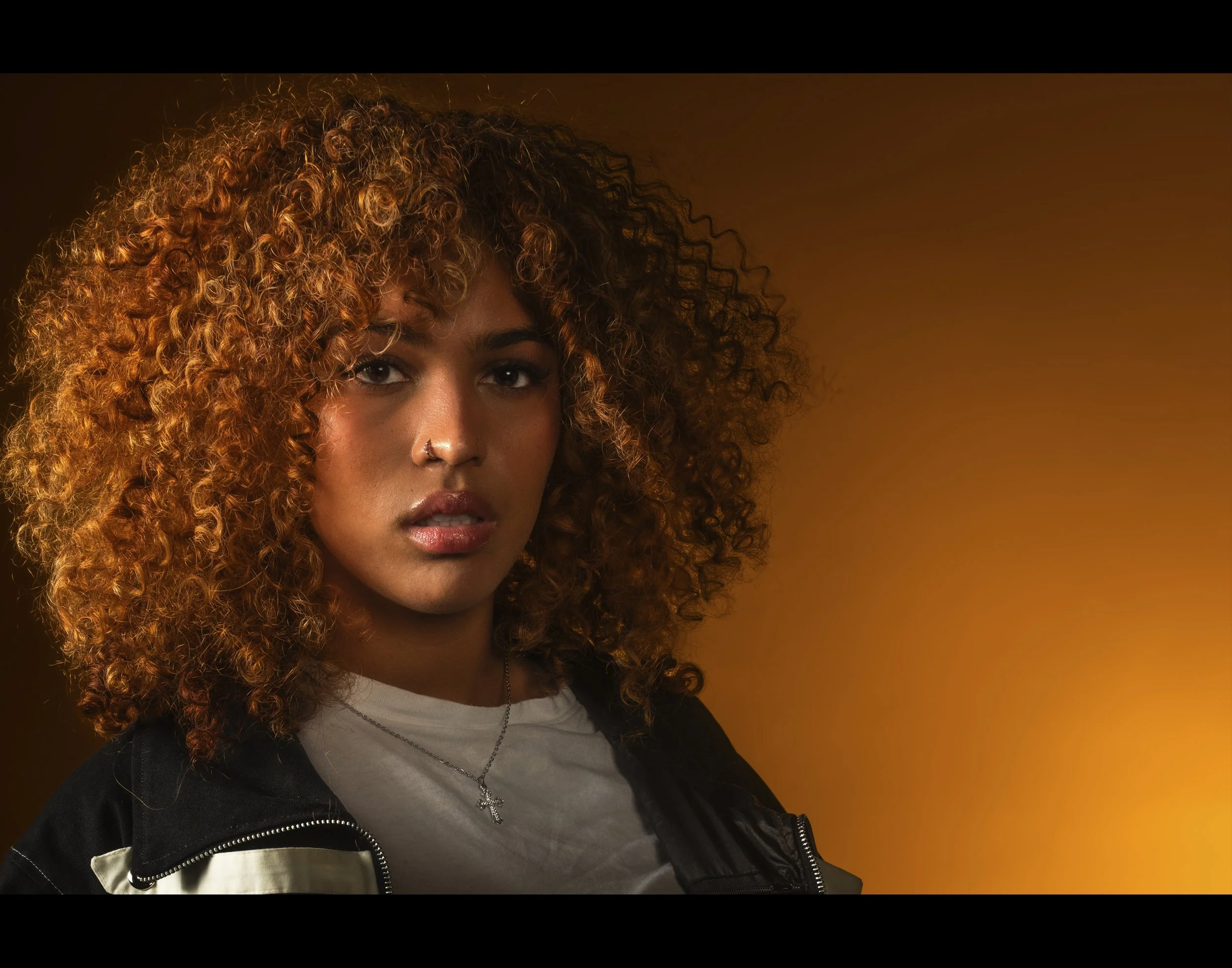 Close-up of a woman with curly red hair wearing a nose ring, necklaces, and a black jacket against an orange background.