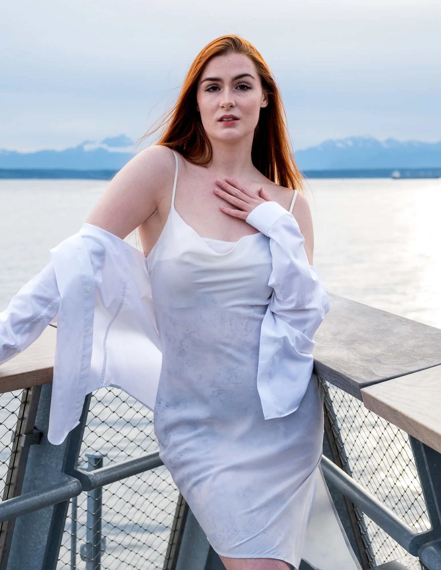 A woman with long red hair standing on a bridge by water, wearing a white dress and loose shirt, looking at the camera with a hand on her chest.