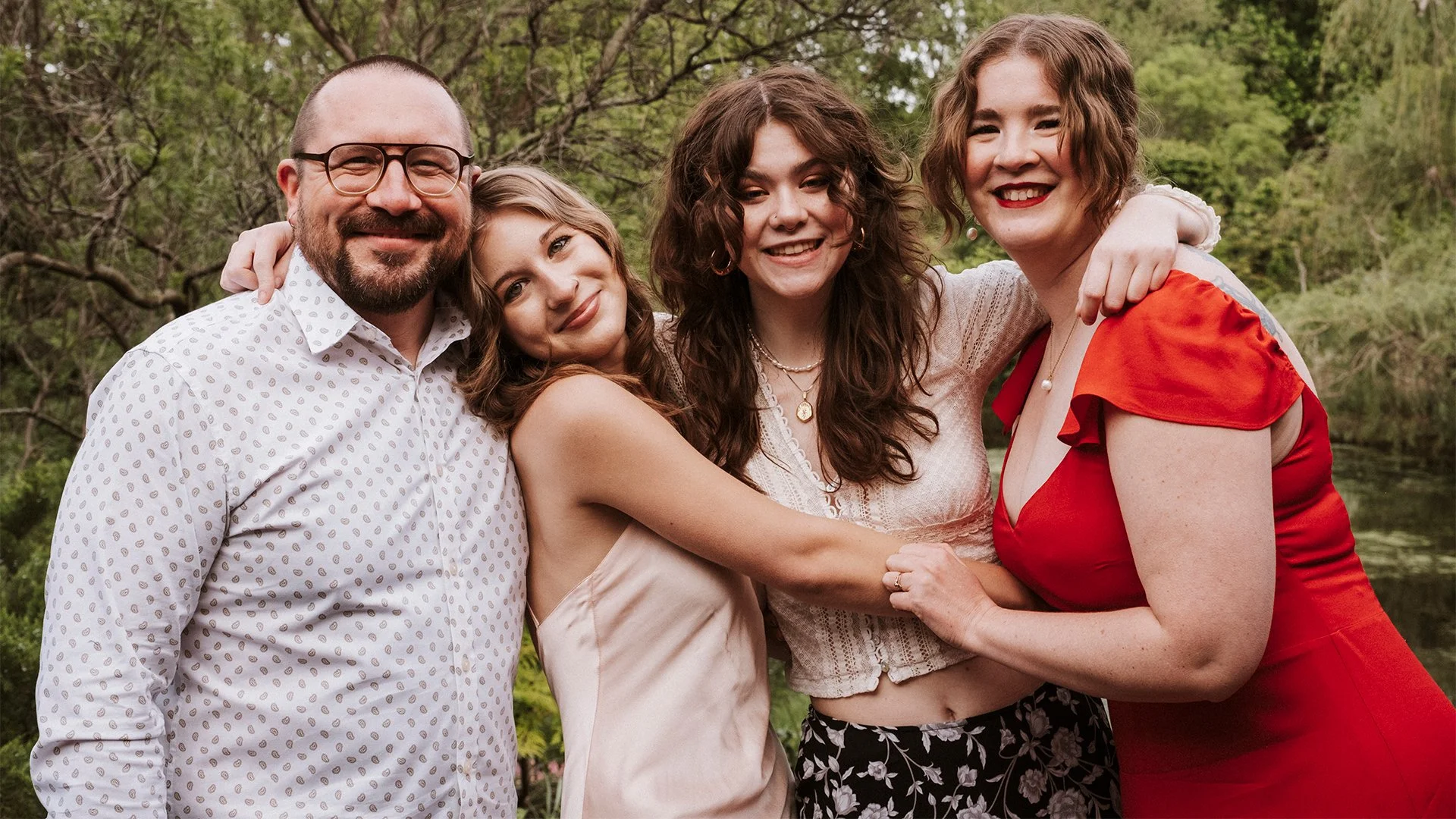 A happy family of five standing close together outdoors in a natural setting, smiling and embracing each other.
