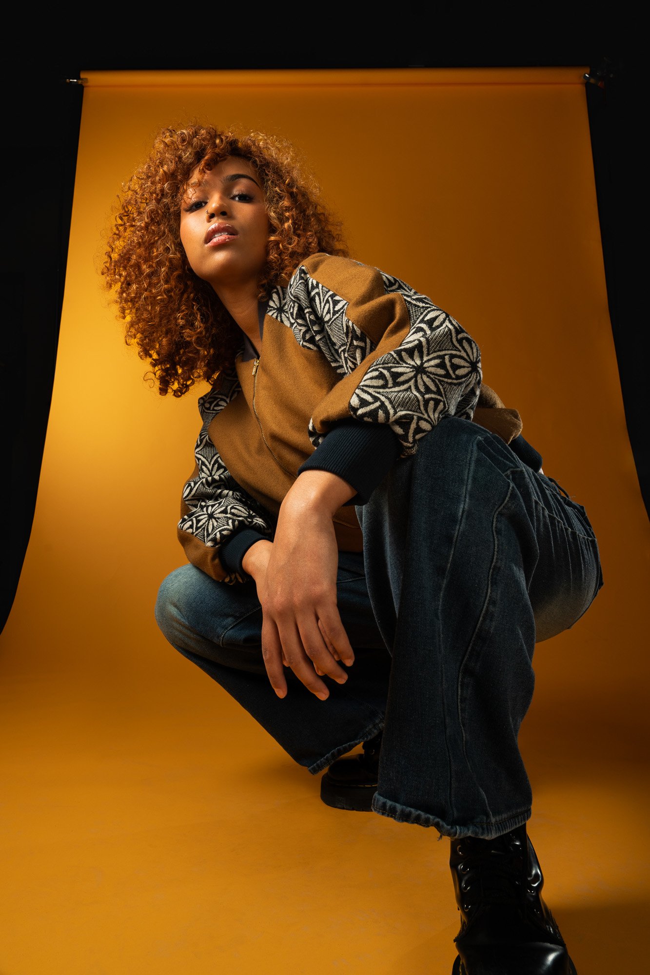 A woman with curly hair and a confident expression is squatting against a yellow backdrop, wearing a patterned jacket, jeans, and black boots.