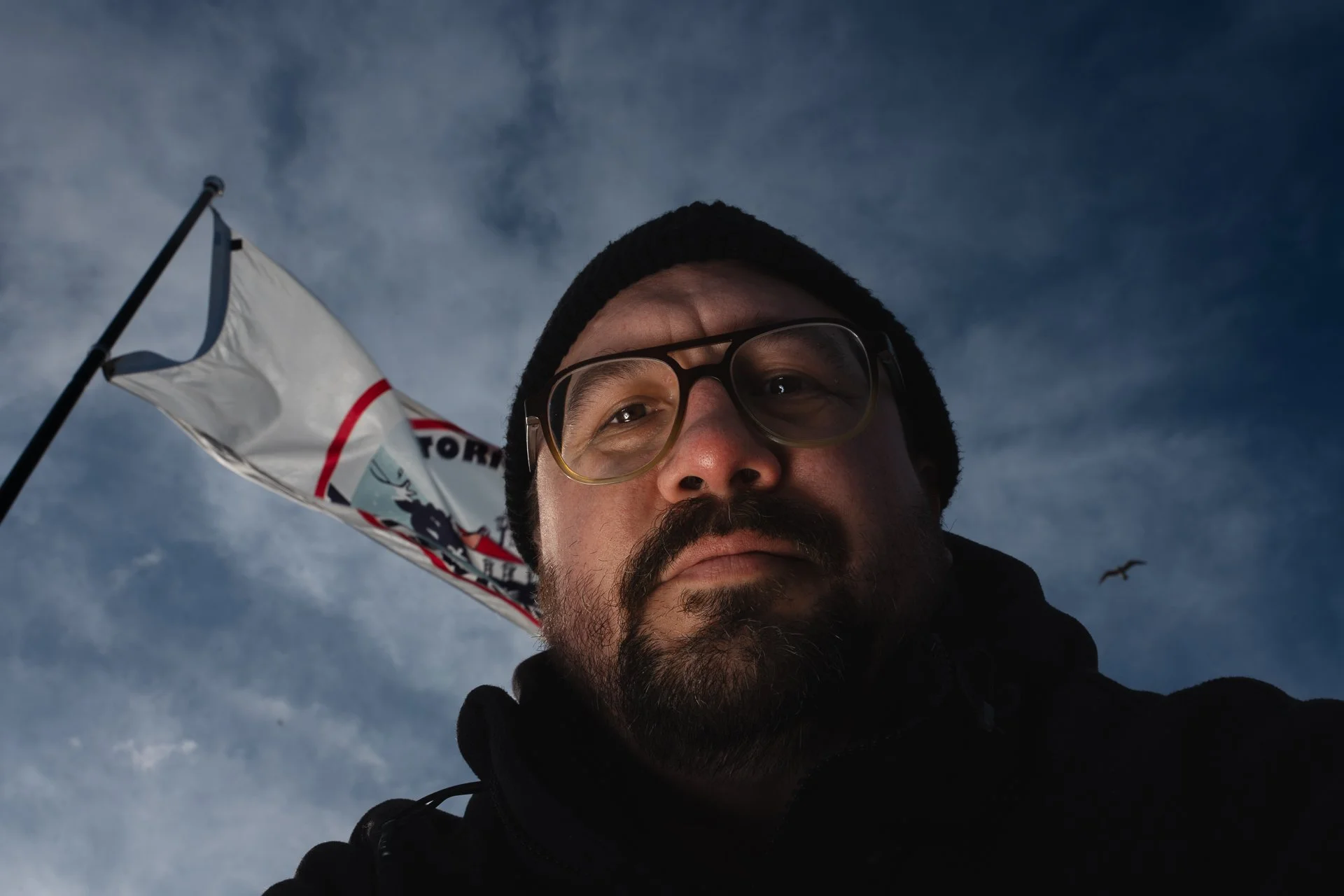 Close-up of a man's face with glasses, wearing a black beanie, outdoor with cloudy sky background, a flag and a bird in flight.