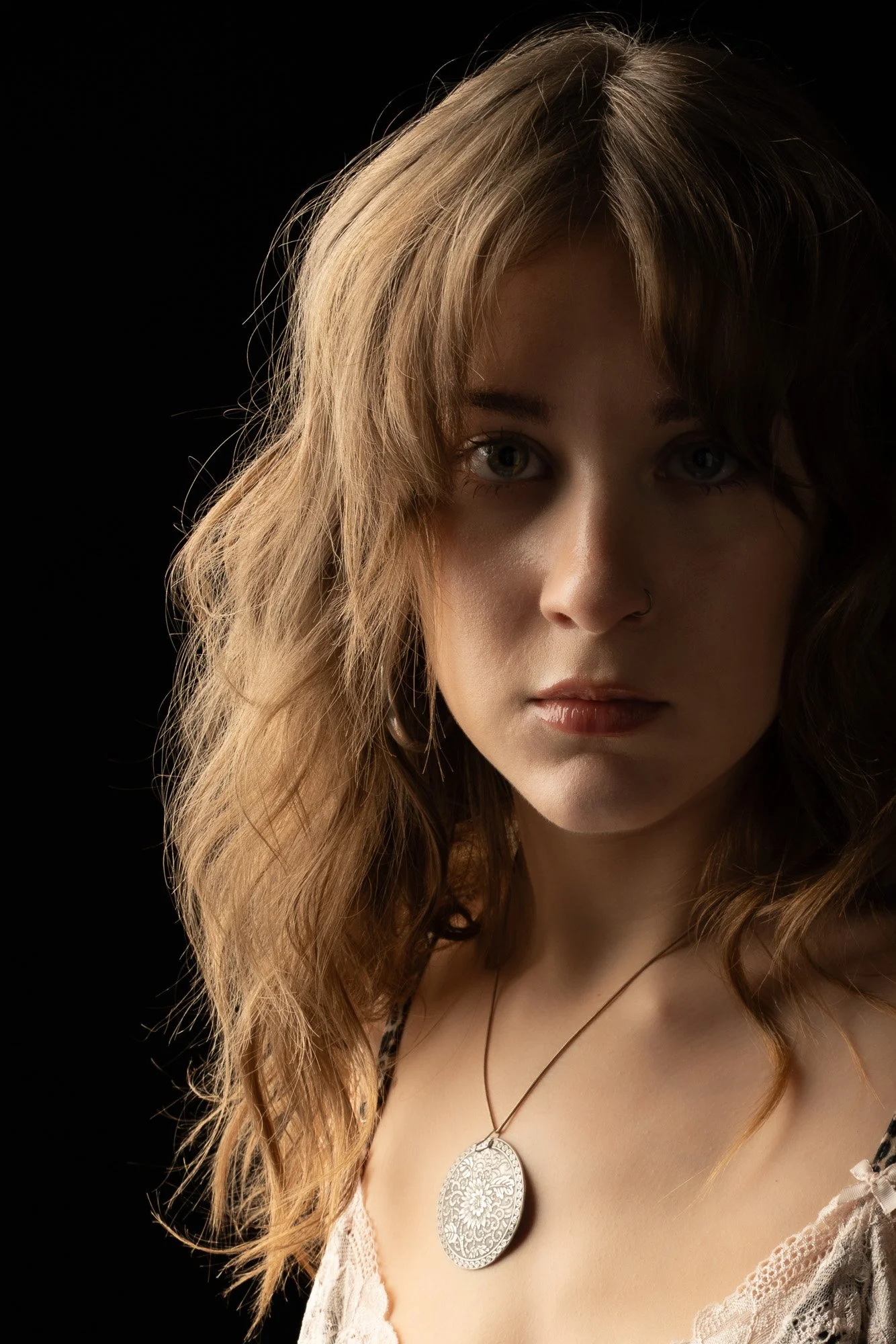 A woman with wavy light brown hair and hazel eyes wearing a lace top and a large round silver pendant necklace, against a black background.