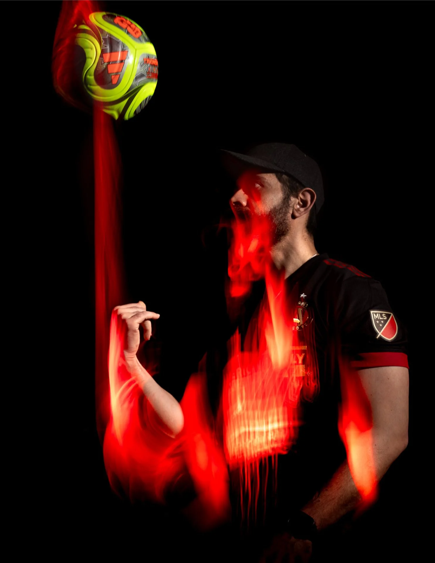 A man wearing a black cap and a soccer jersey with MLS logo, holding a colorful soccer ball, with red light streaks over the image.