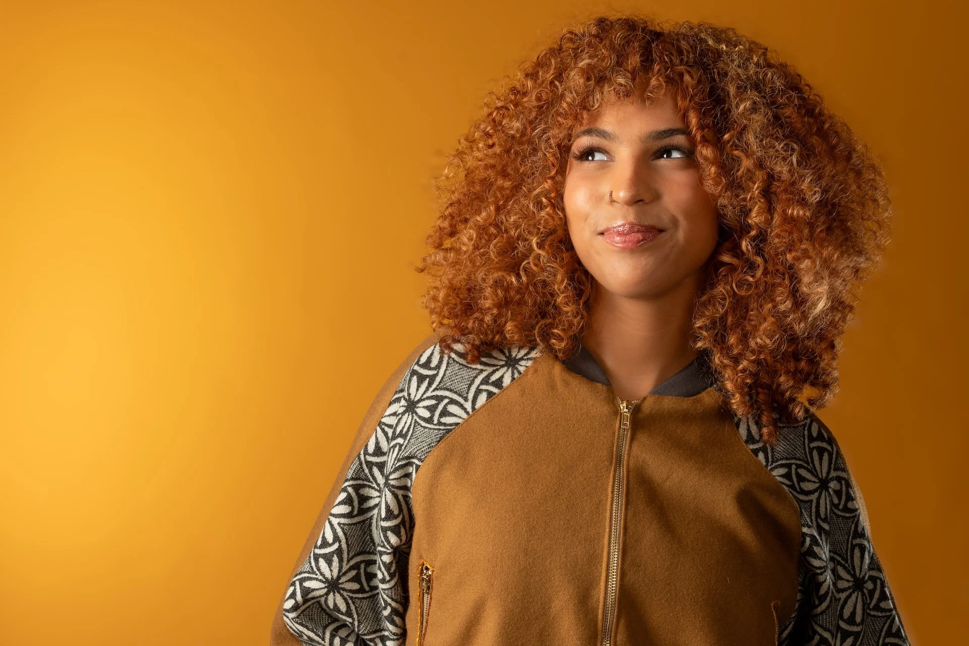 A woman with curly red hair and a nose piercing looking to her left, wearing a brown jacket with a zipper and patterned sleeves, against a solid yellow background.