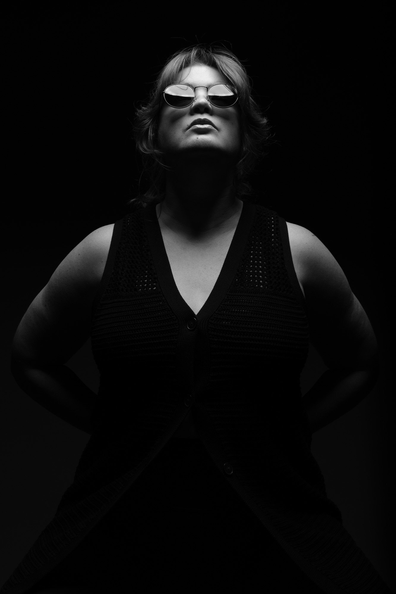 Black and white portrait of a woman wearing sunglasses, standing with hands on hips, against a dark background.