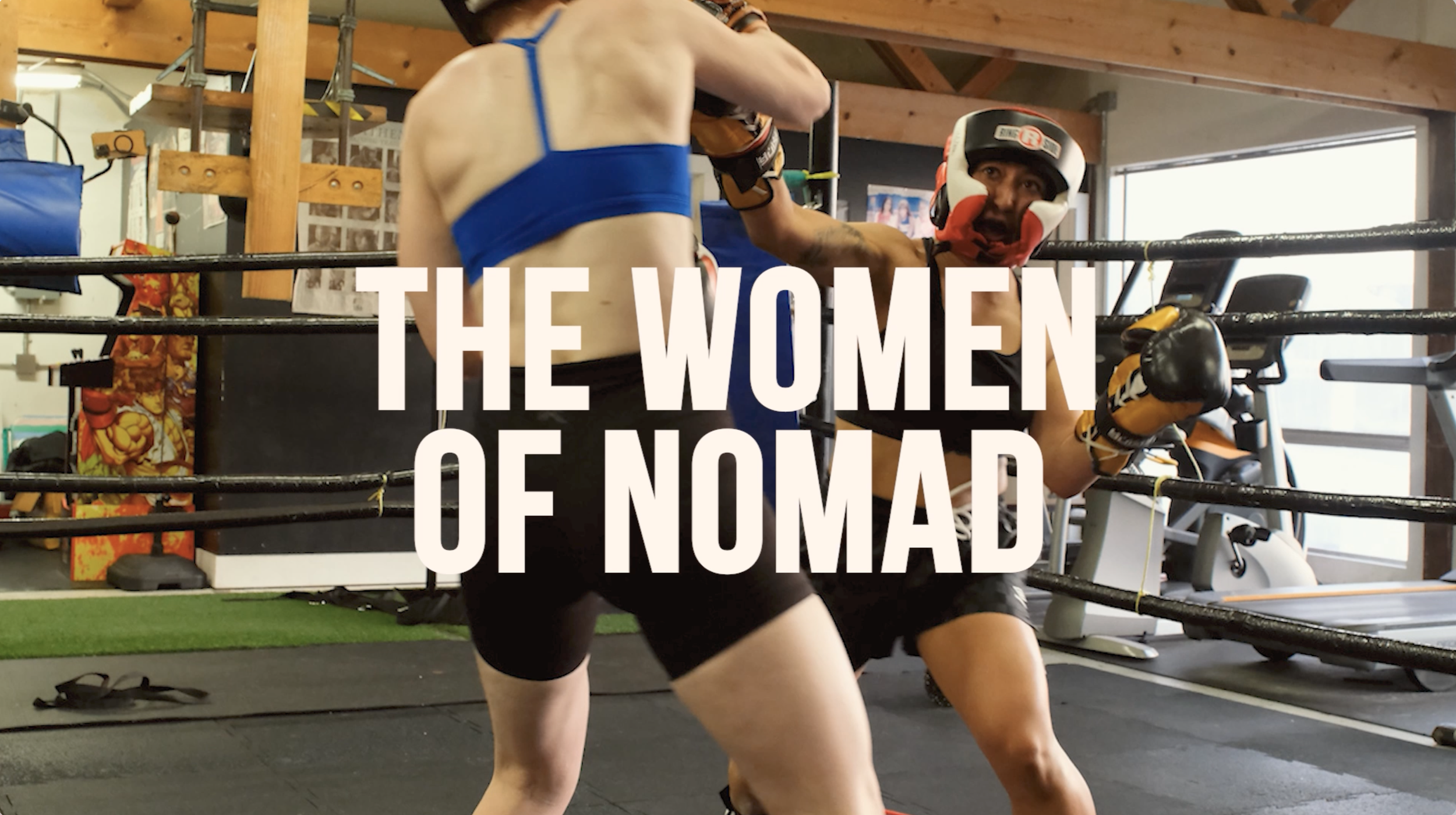 Two women fighting in a boxing ring while wearing protective headgear and gloves. One woman is throwing a punch at the other, who is leaning back. The scene is set in a gym with wooden beams, exercise equipment, and a colorful poster of a superhero on the wall.