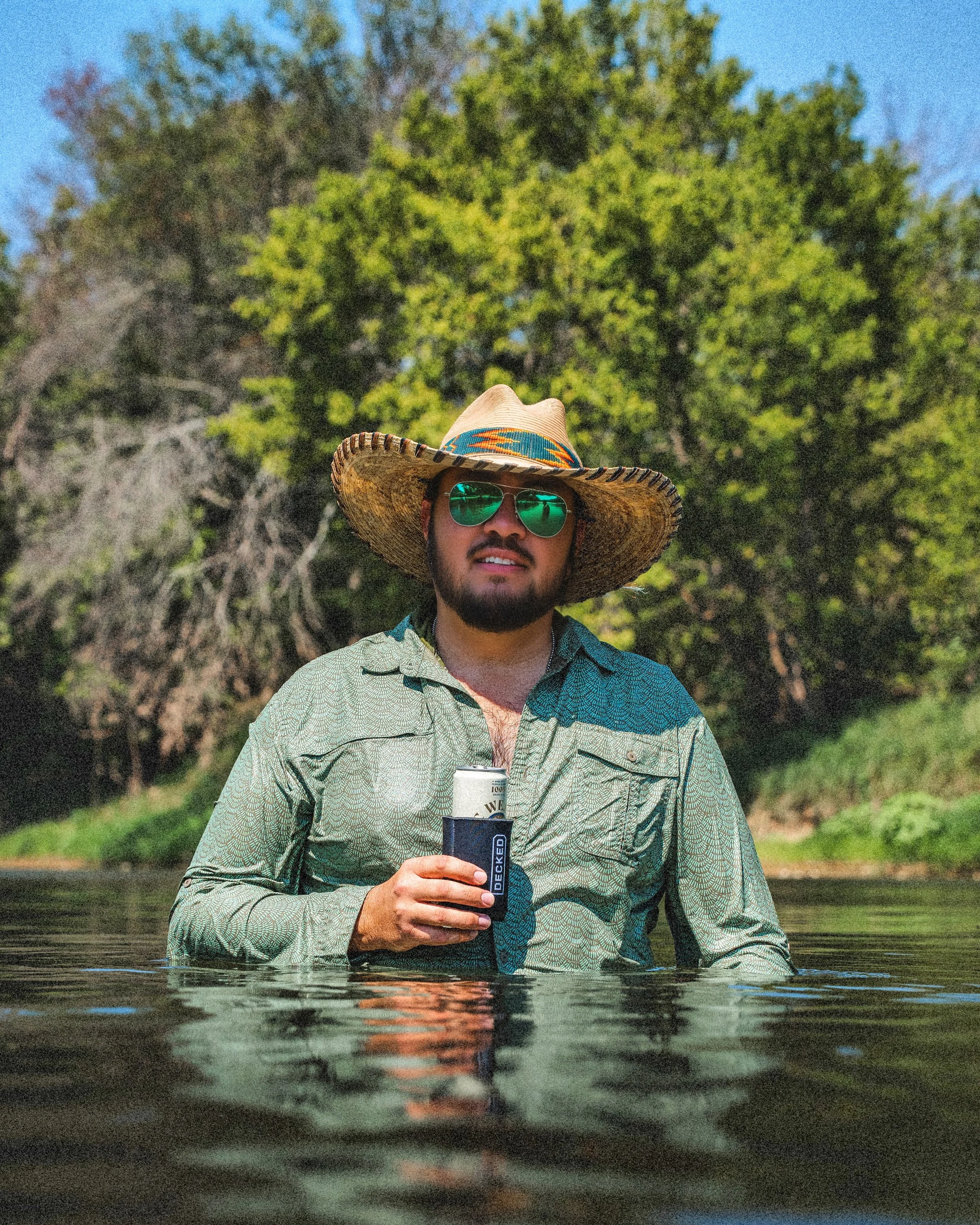 A man wearing a wide-brimmed straw hat and green reflective sunglasses standing in water, holding a cocktail. He is dressed in a patterned, button-up shirt and is surrounded by a lush, green forest with a large tree and blue sky in the background.