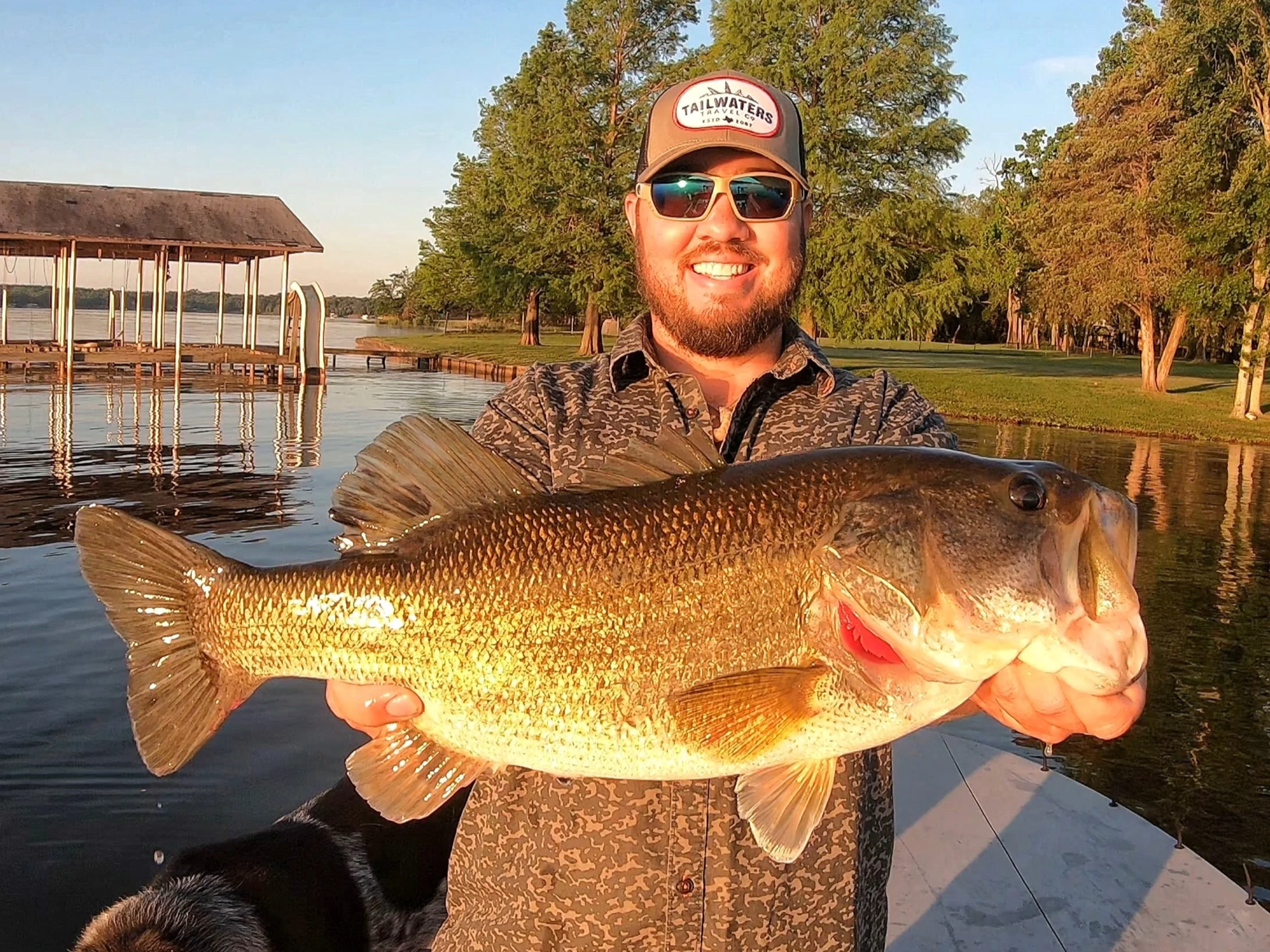 A man wearing sunglasses and a baseball cap labeled 'Tailwaters Travel Co' is smiling and holding a large fish while standing near a body of water, with green trees and a dock visible in the background.