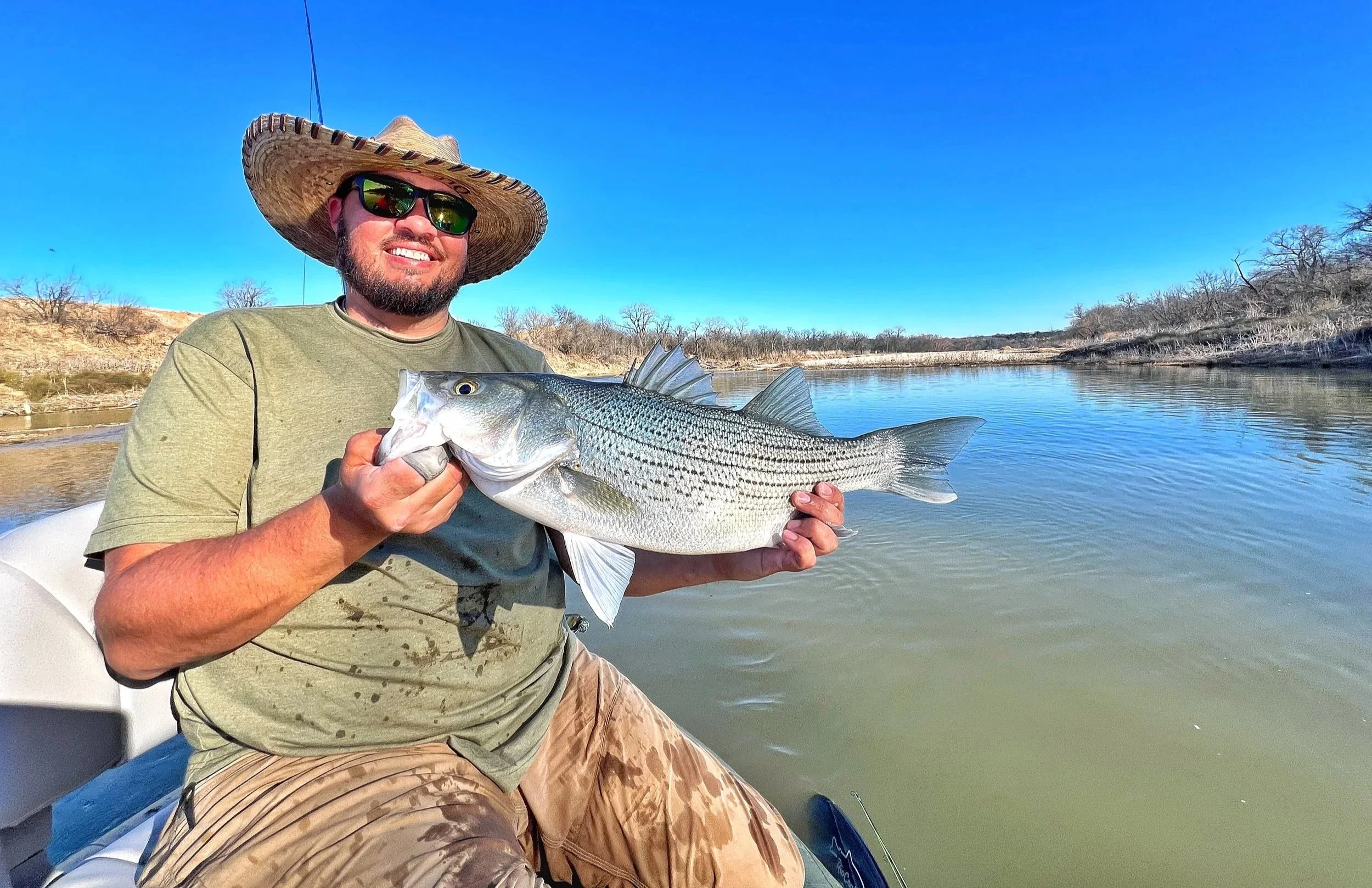A man wearing a straw hat and sunglasses while sitting in a boat, holding a large fish he caught, with a river and barren trees in the background on a sunny day.