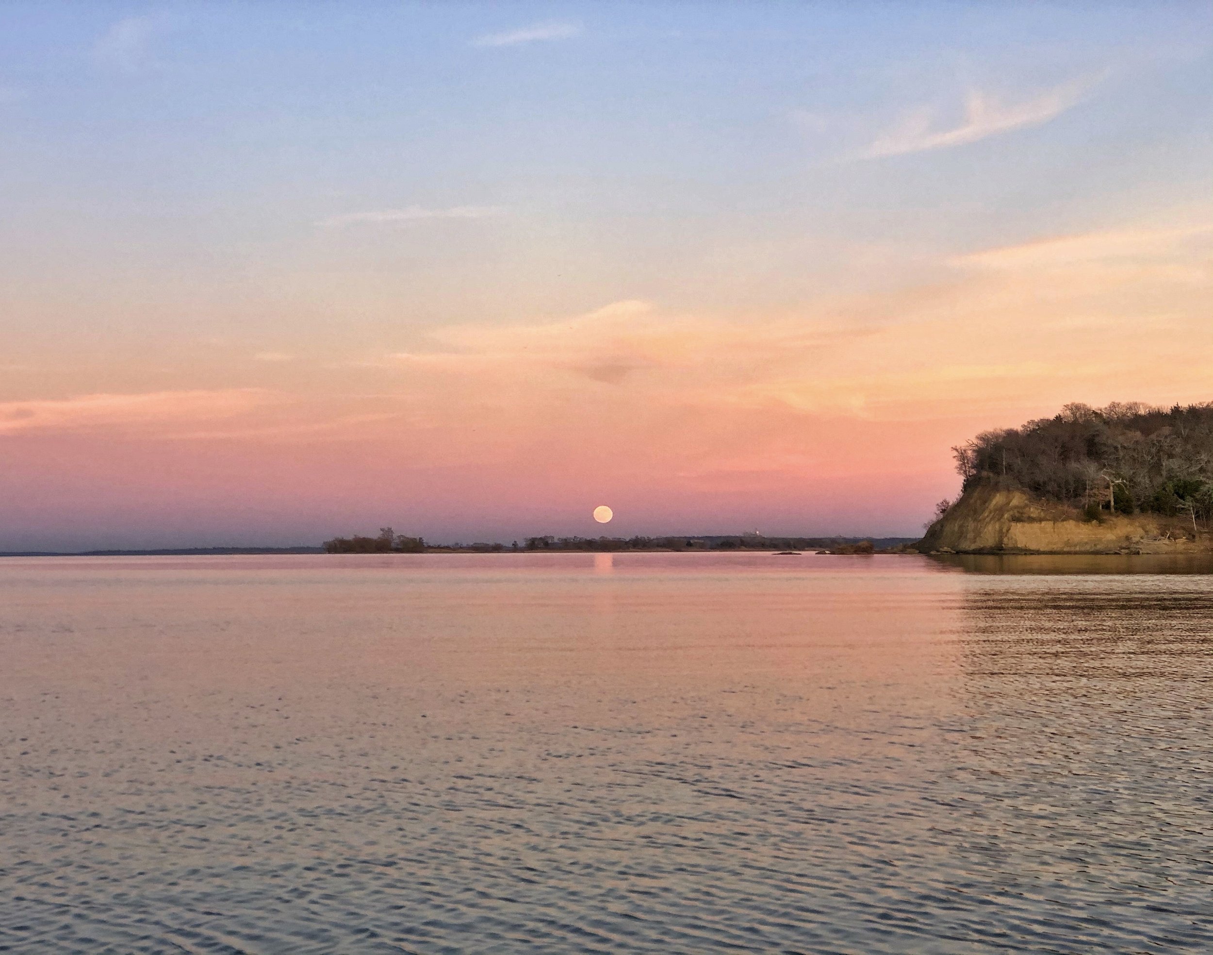 A peaceful scene of a pastel-colored sky at sunset with a full moon rising over a body of water with a hill covered in trees on the right side.