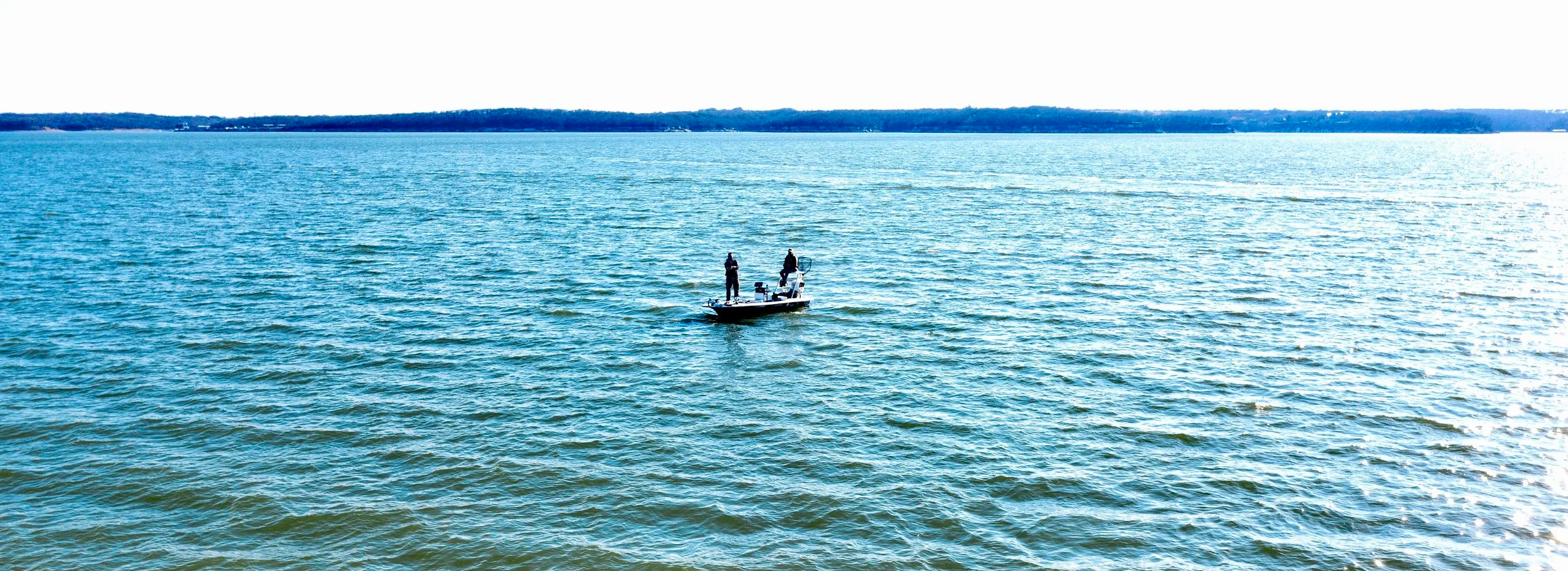 Two people fishing on a skiff in a large body of water with a distant shoreline under a clear sky.