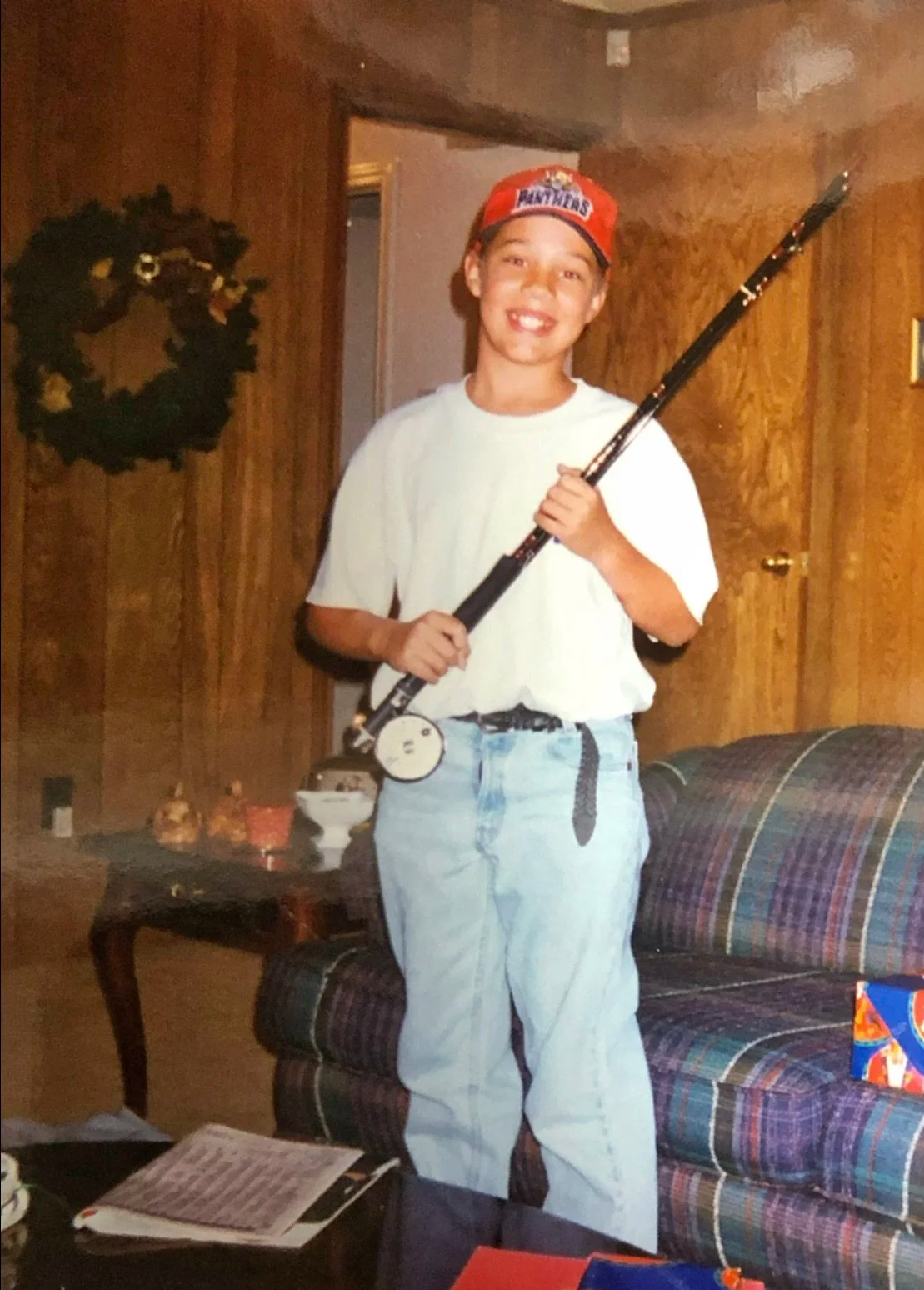 A boy smiling and holding a fishing rod inside a room with wood-paneled walls, a plaid couch, and a side table with snacks and bowls, wearing a red cap and casual clothes.