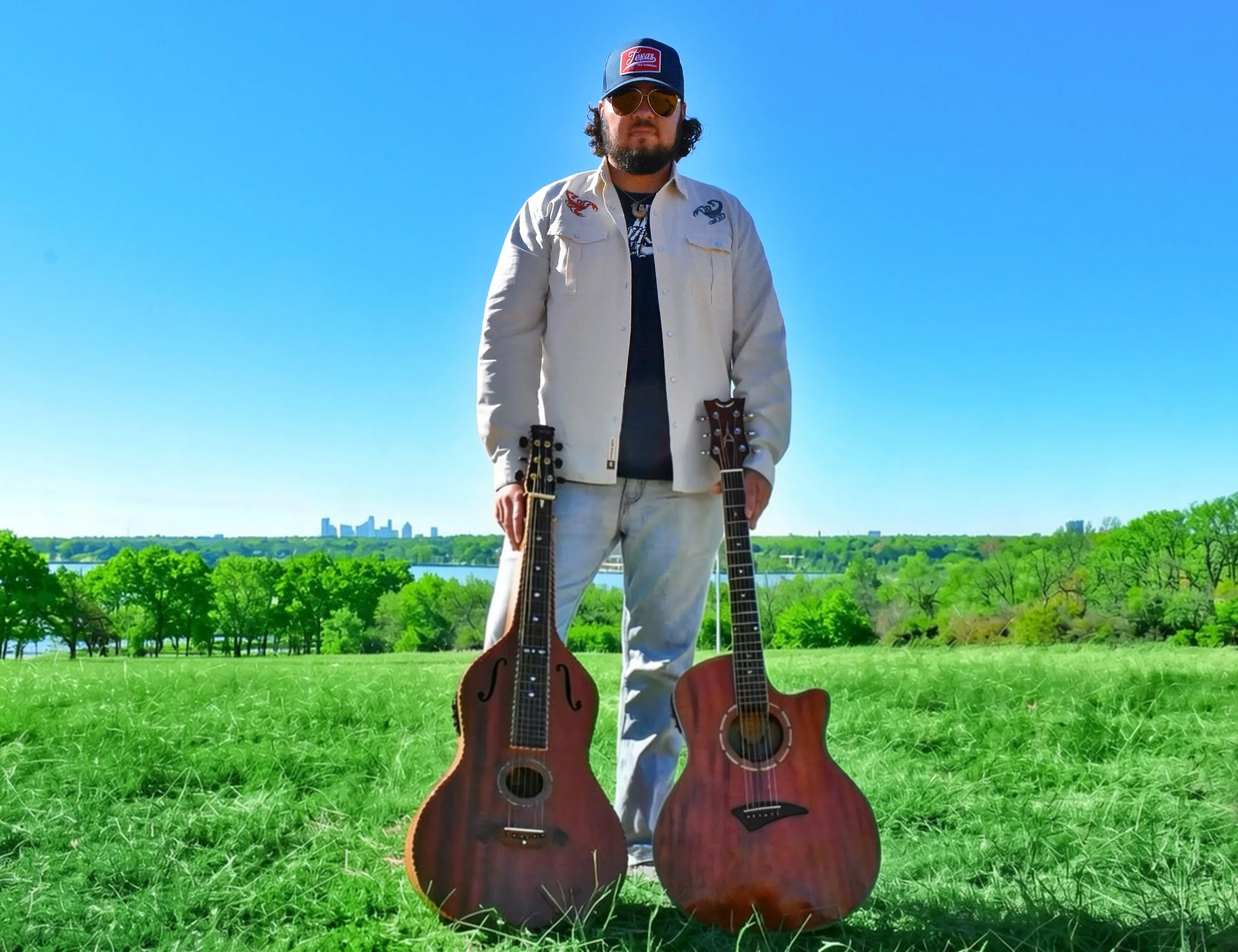A man with sunglasses and a baseball cap standing in a grassy outdoor area with two acoustic  guitars on the ground, with a city skyline and blue sky in the background.