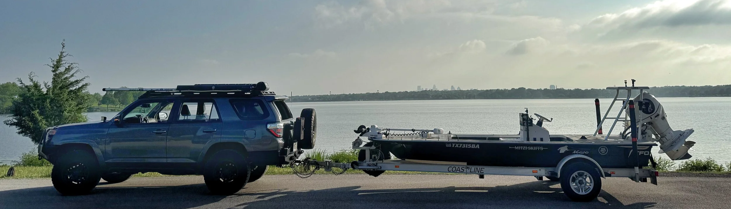 A blue SUV parked by a lake on a road, towing a boat on a trailer. The boat has a white outboard motor, and the scene is set during the day with a partly cloudy sky and distant city skyline.