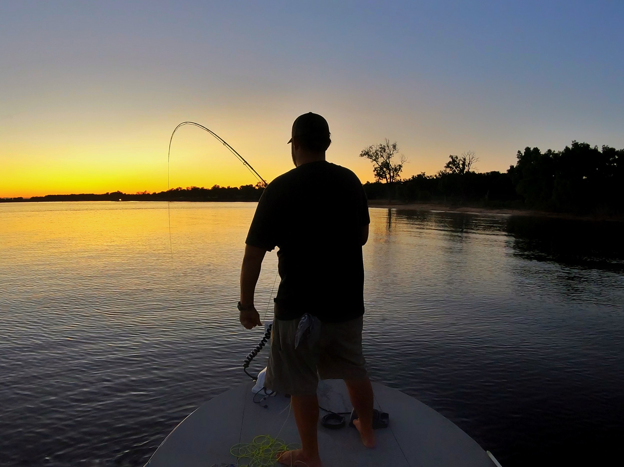 A silhouette of a man fishing off a boat at sunset on a calm river with trees in the background.