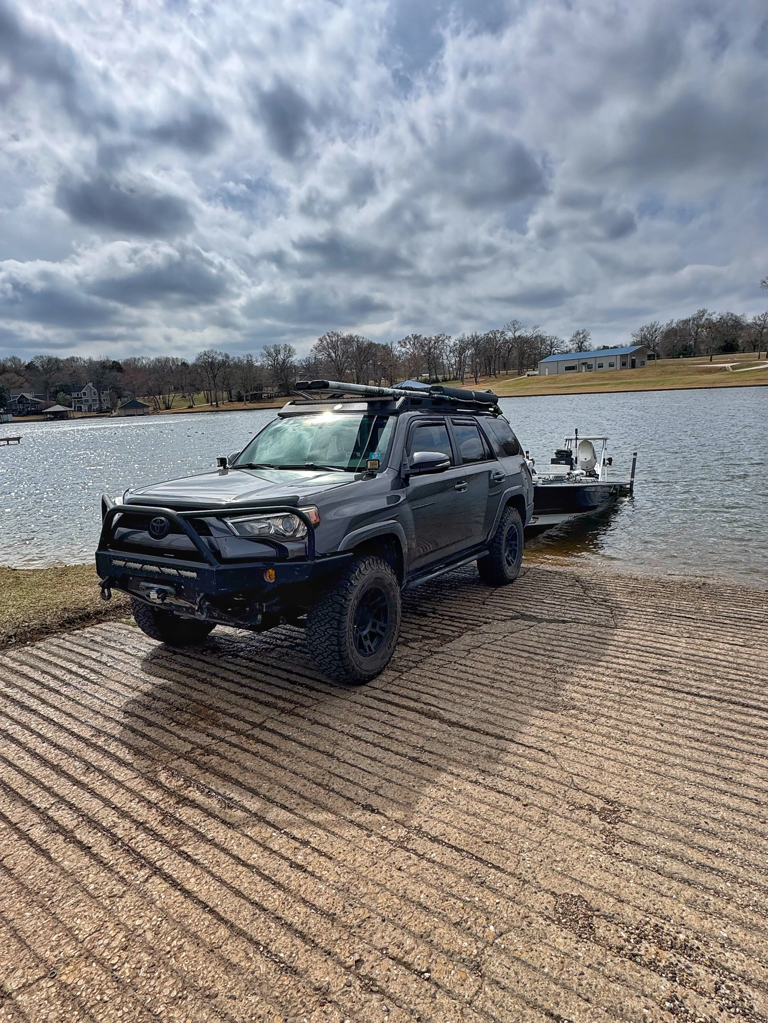 A gray Toyota 4x4 pickup truck with a roof rack and a front bumper guard, attached to a boat, parked on a concrete boat ramp at a lake. The background shows houses, a grassy area, and leafless trees under a cloudy sky.