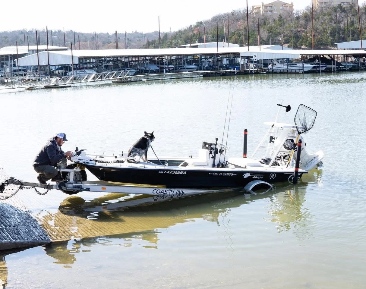 A man and a dog on a fishing boat docked at a marina, with boats and covered slips in the background.