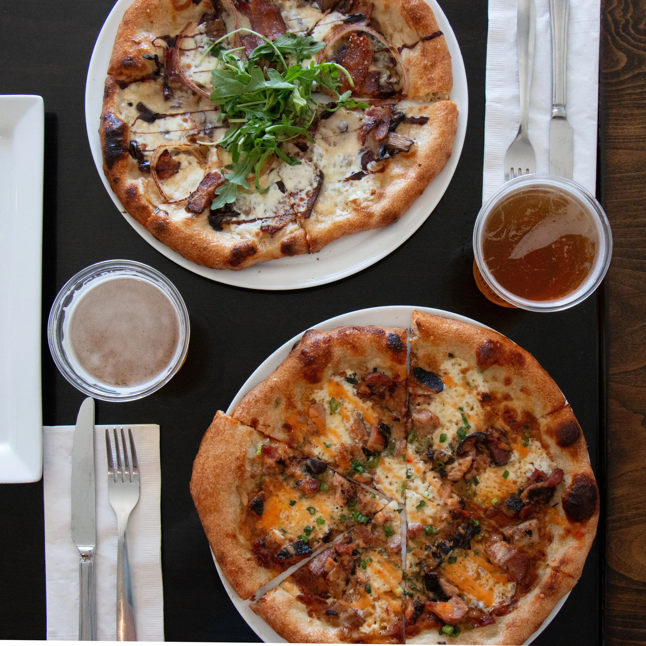 A white plate of pizza topped with arugula, bacon, mushrooms, cheese, and green onions. There are two cups of tea or coffee, a fork, a knife, and a spoon on a napkin.