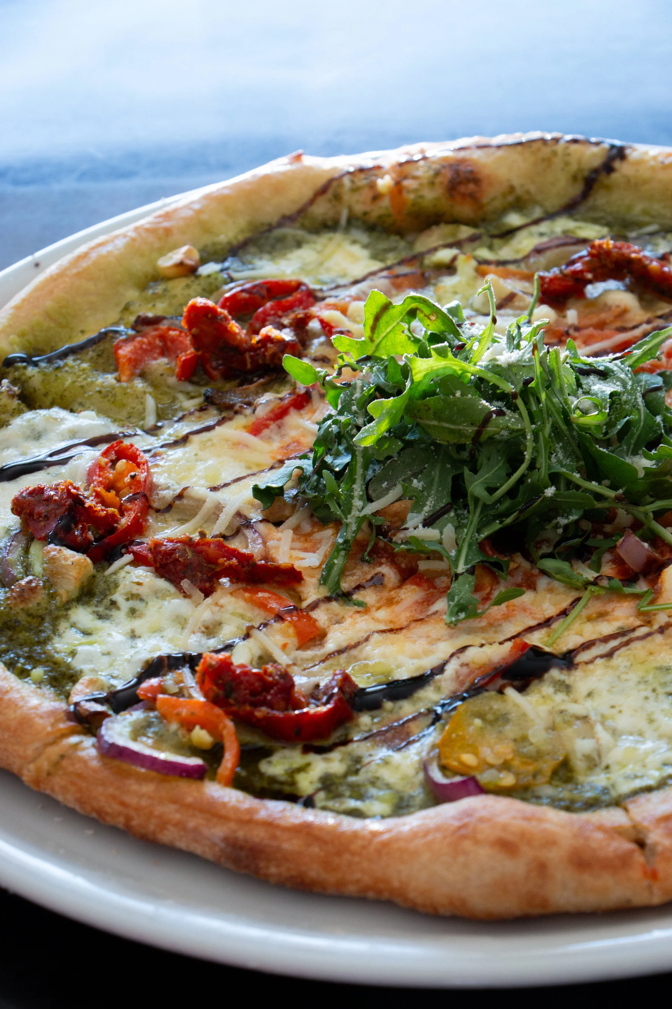 A pizza topped with arugula, tomatoes, onions, cheese, and drizzled with balsamic glaze on a white plate.