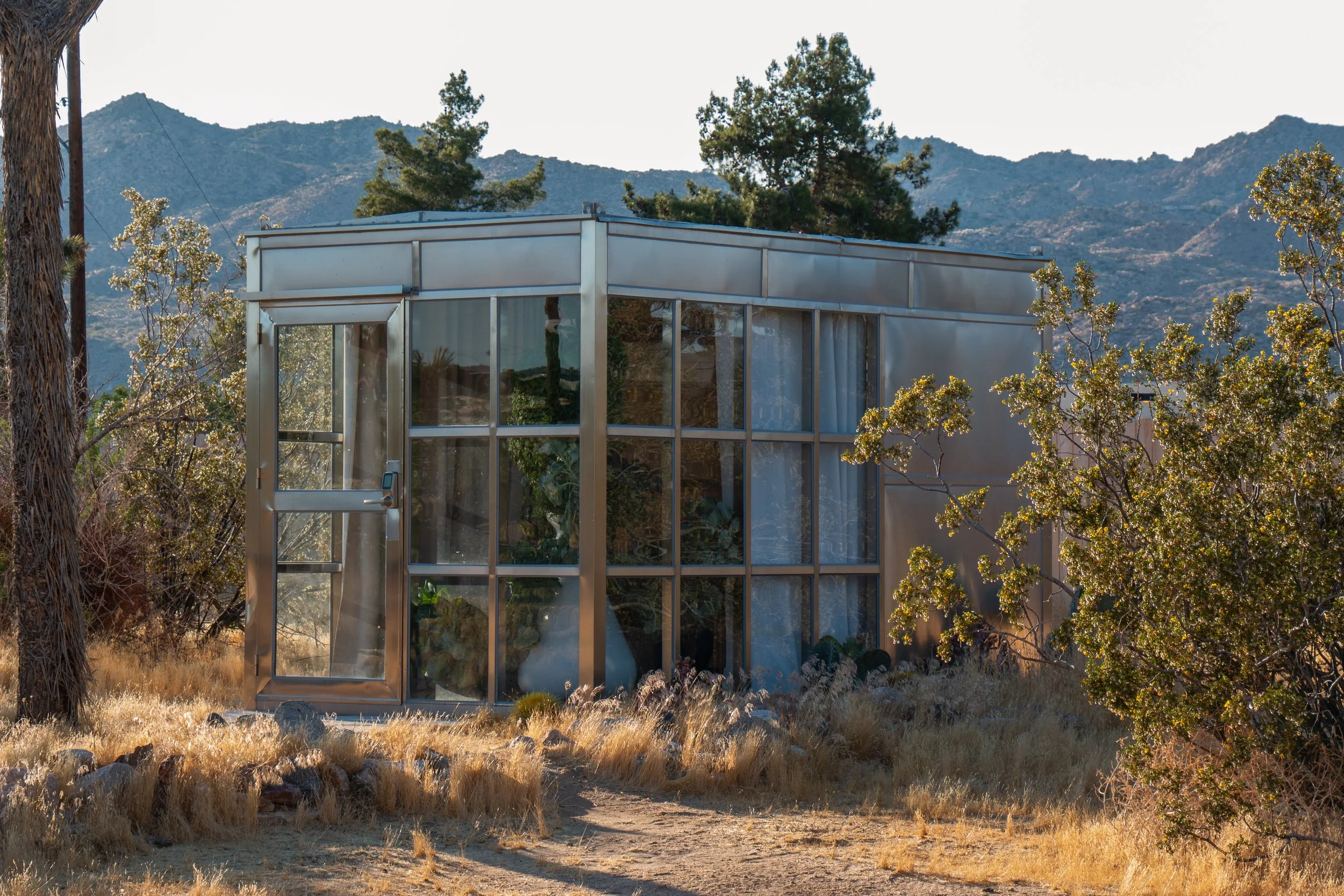 A modern glass house in a desert landscape with dry grass and rocks, surrounded by trees, with mountains in the background.