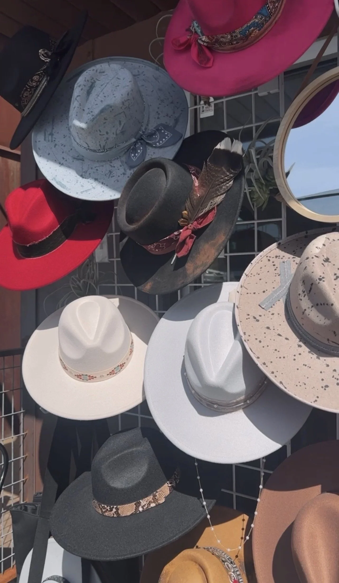 A display of various hats, including wide-brimmed, fedora, cowboy hats, and chunky knit styles, arranged on a tiered rack with sunlight casting shadows.