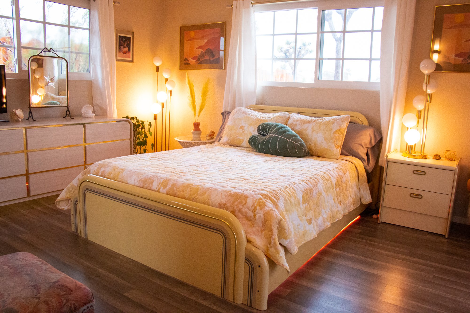 A cozy bedroom with a bed, large windows, and decorative lighting. The bed has light-colored, textured bedding and pillows, including a dark green leaf-shaped pillow. Next to the bed is a nightstand with a modern lamp, and there are framed artwork an