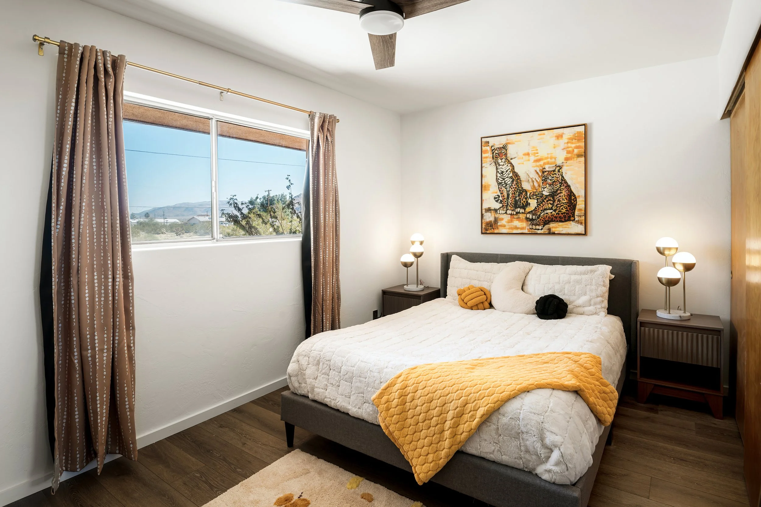 A bedroom with a large window, white walls, and a ceiling fan. The bed has a white quilt, orange blanket, and pillows, with two small nightstands, each holding a modern lamp. A colorful painting of two leopards is on the wall above the bed. The room 
