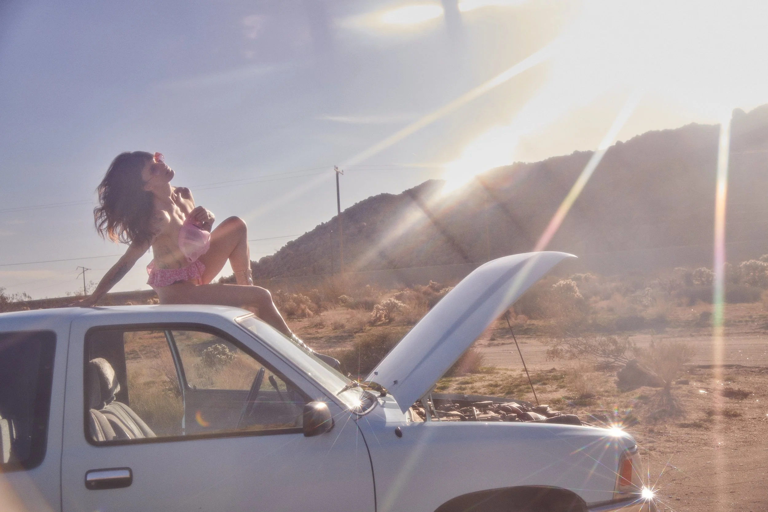 A woman sitting on the roof of a white pickup truck with the hood open, in a desert landscape, with mountains and power lines in the background during sunset or sunrise.