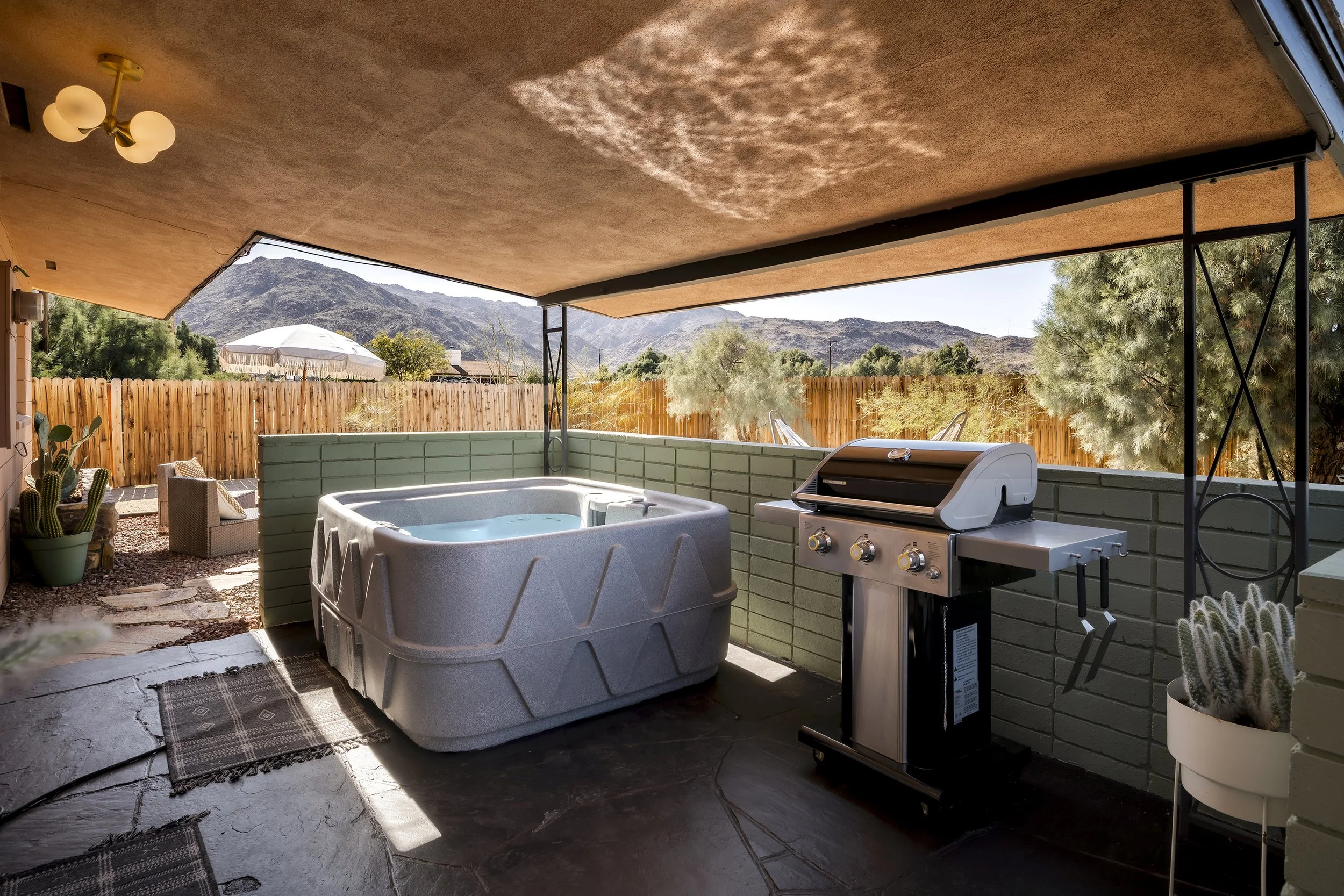 Covered patio area with a hot tub, a barbecue grill, potted cactus plants, outdoor seating, and mountain views in the background.