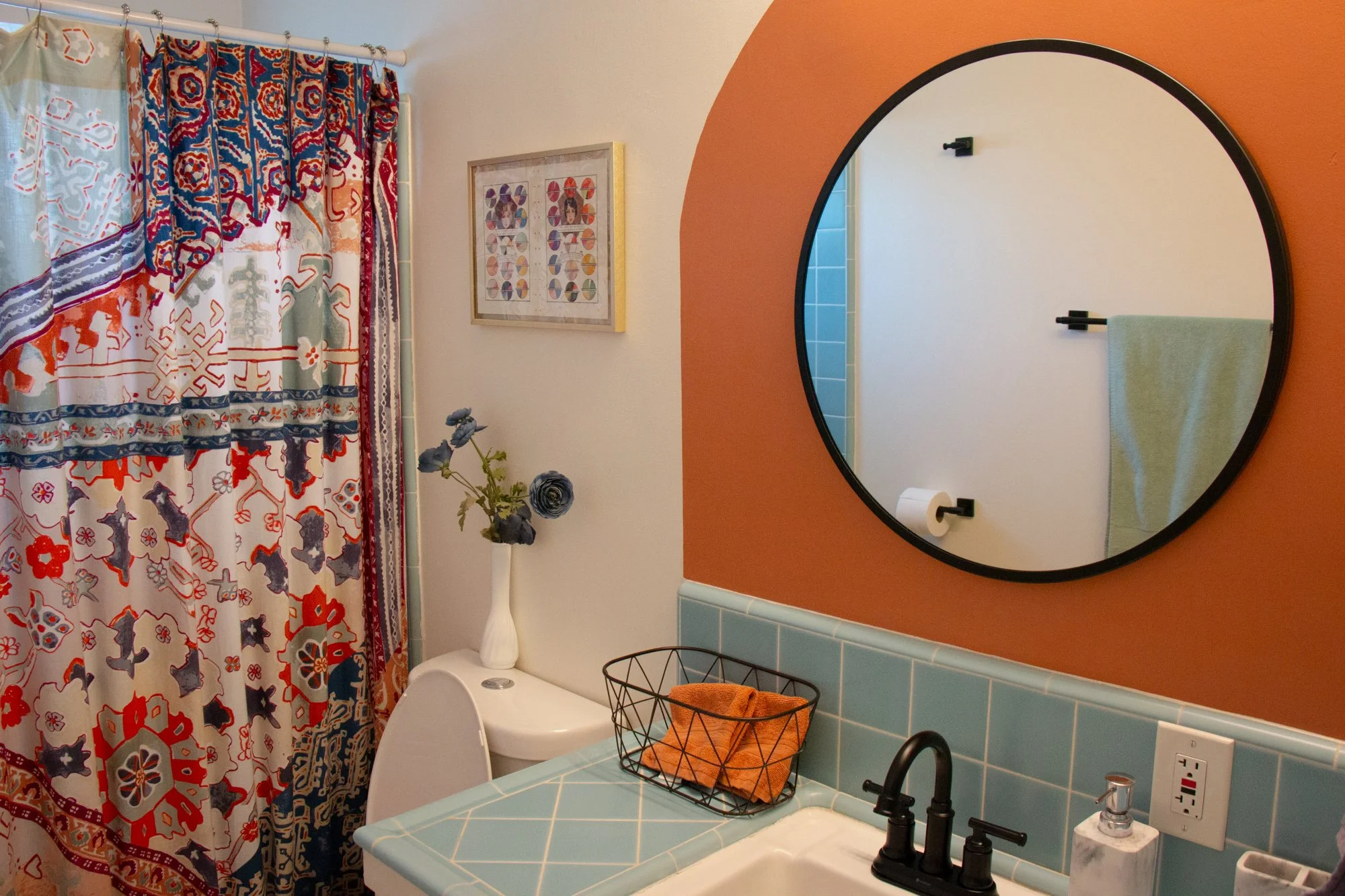 Bathroom with a colorful patterned shower curtain, blue tiled countertop, black faucet, round mirror on orange wall, small framed artwork, white vase with purple flowers, and a wire basket with orange towels.