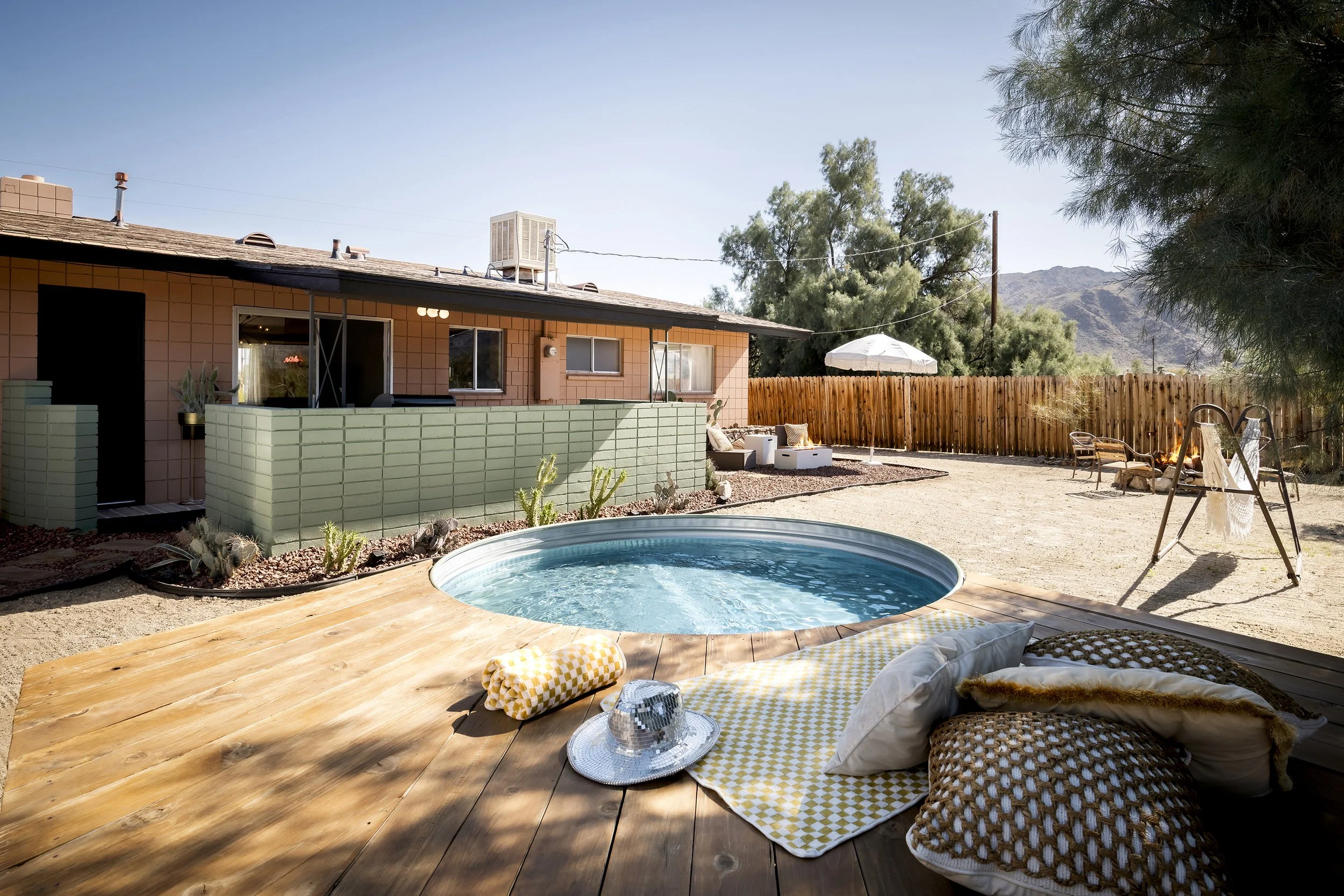 A backyard area with a small swimming pool in the foreground, yellow and white checkered towels and pillows on a wooden deck, a house with a pink exterior and green low wall, patio furniture with cushions, a white umbrella, trees, a mountain in the b