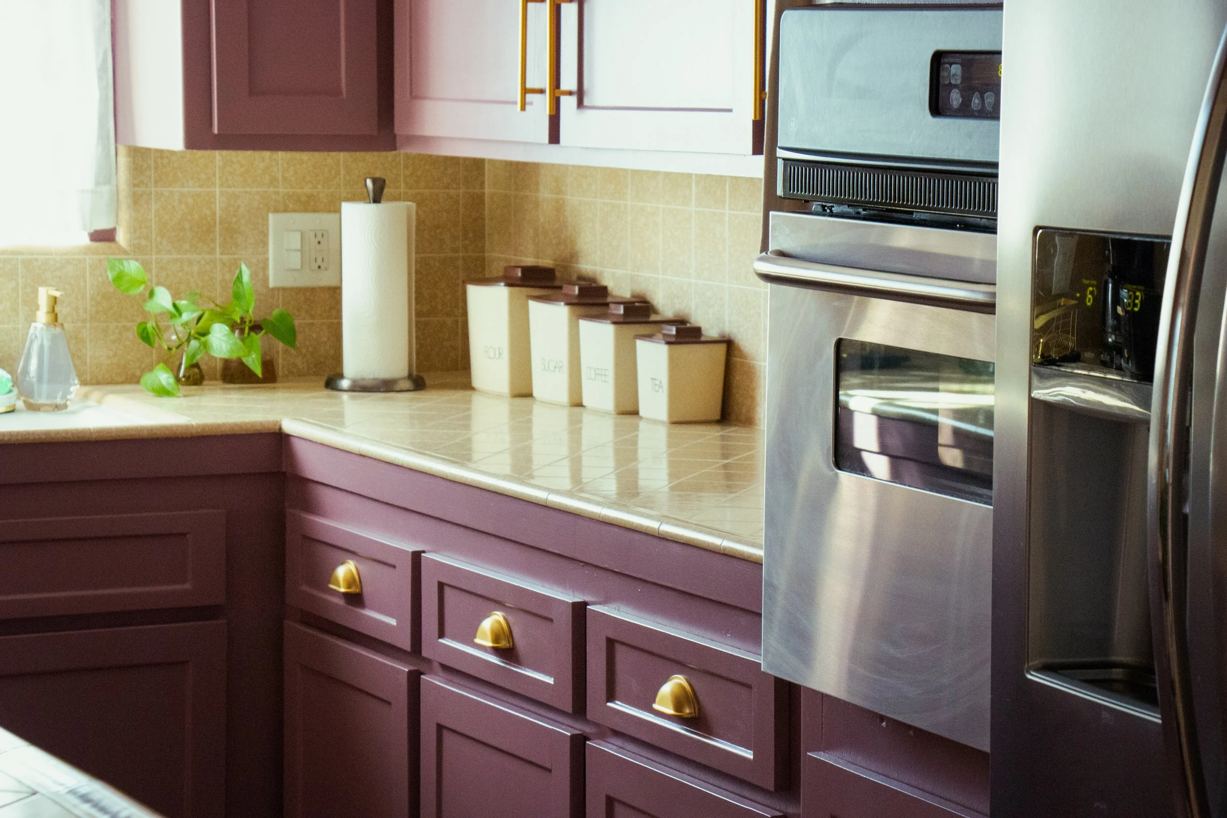 Kitchen with purple cabinets, beige tiled backsplash, stainless steel oven, and white canisters labeled flour, sugar, coffee, and tea.