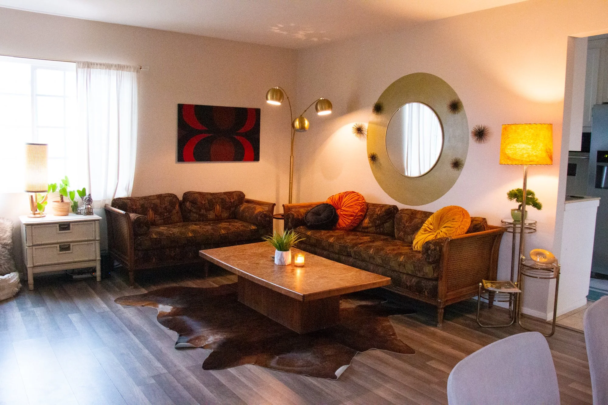 Living room with patterned sofa, round wooden coffee table, large window with white curtains, decorative wall mirror with starburst design, abstract wall art, floor lamp with brass finish, yellow lampshade, potted plants, small side tables, and hardw
