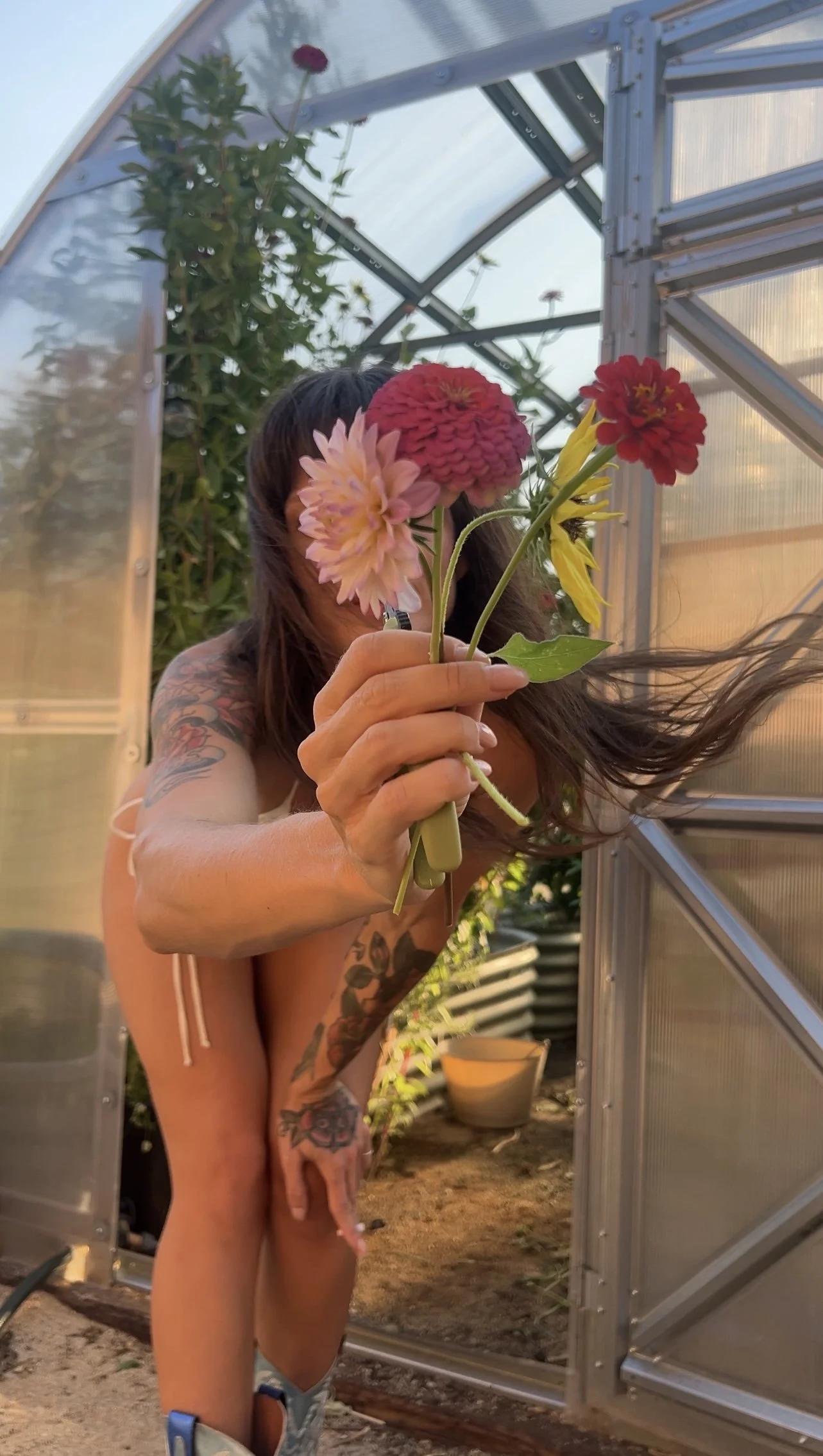 A woman with tattoos holding a colorful bouquet of flowers in a greenhouse.