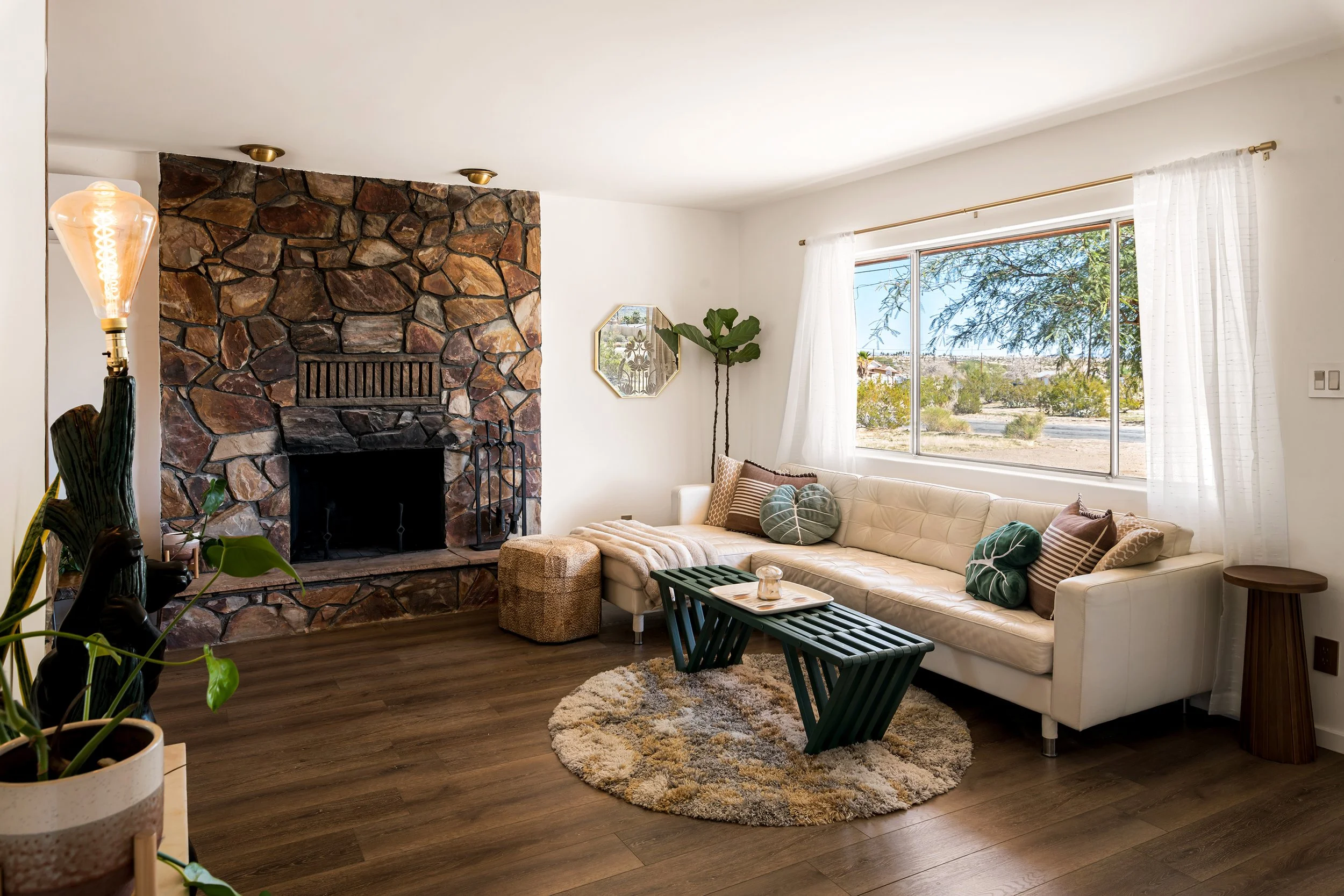 Living room with a stone fireplace, large window, cream sectional sofa with pillows, green bench, round rug, and indoor plants.