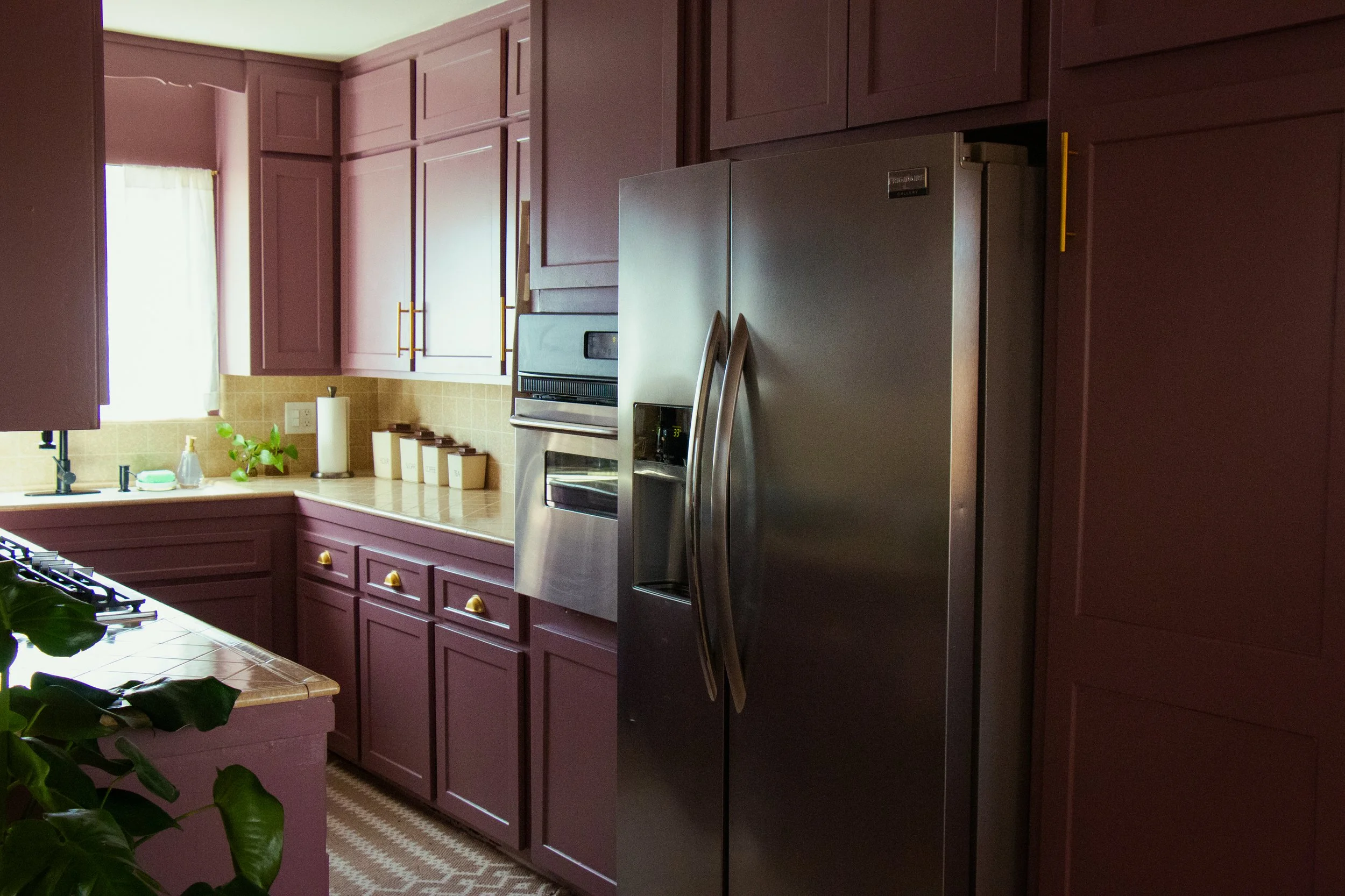 A kitchen with purple cabinets, stainless steel refrigerator and oven, beige tiled backsplash, and a window with white curtains.